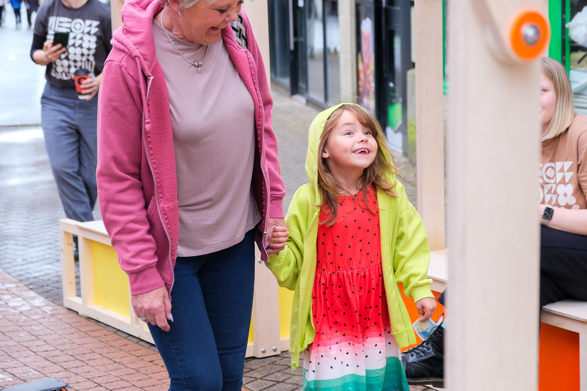 A smiling child in a colourful dress and green hoodie holds hands with an older woman in a pink jacket, walking outside under an arch.