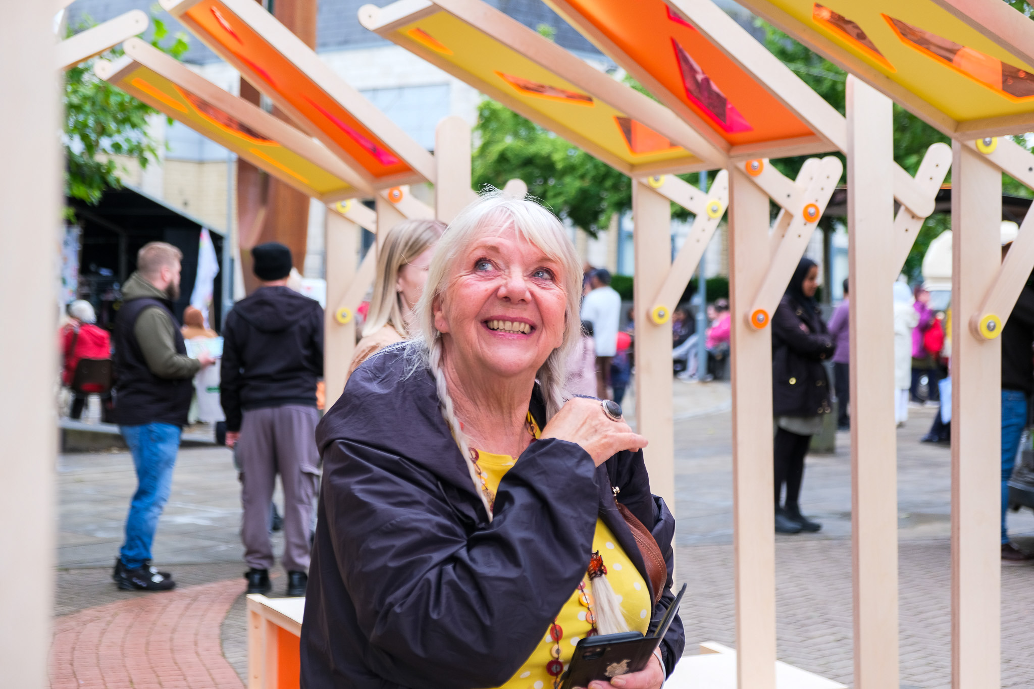 An older woman with grey hair smiles whilst sitting under a colourful wooden structure at an outdoor event. People and trees are visible in the background, creating a lively atmosphere.