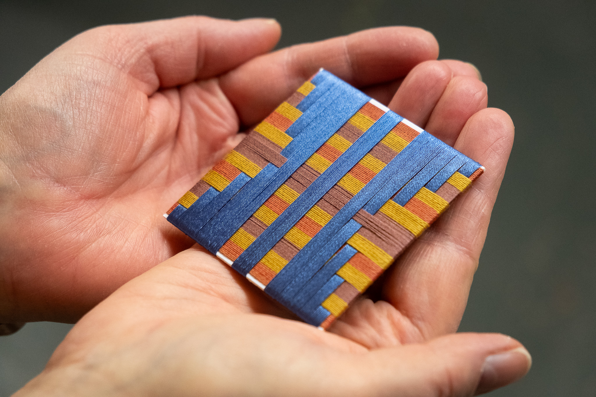 A person holds a small, square, colourful object made of interwoven strips in blue, orange, yellow, and brown, pattern resembling a mosaic or textile art piece.