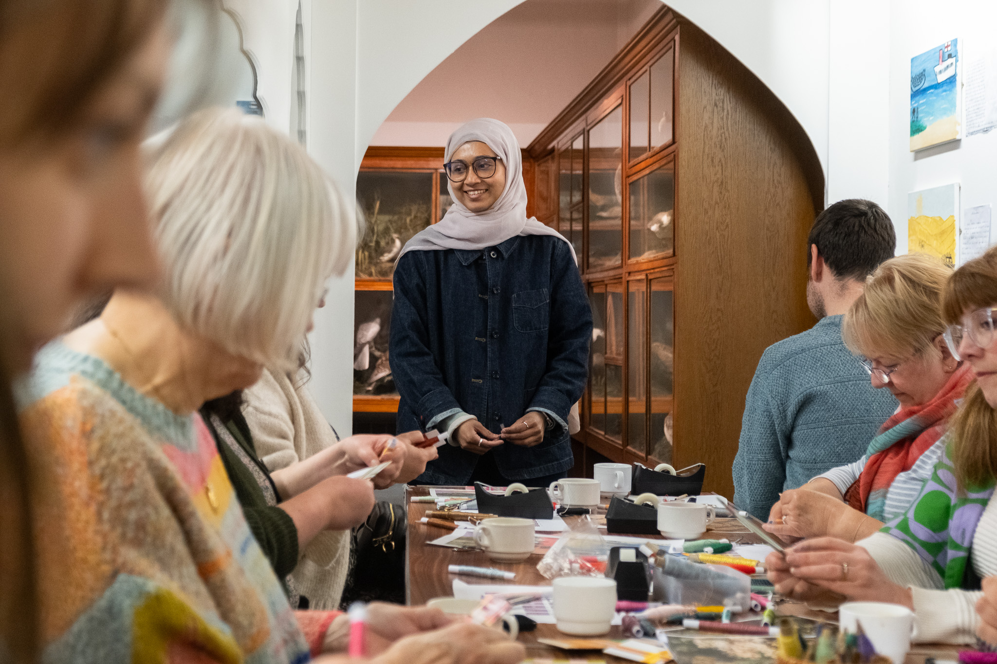 A woman in a hijab and glasses stands smiling at the front of a table where several people are seated, working on arts and crafts with paper, markers, and cups in a brightly lit room.