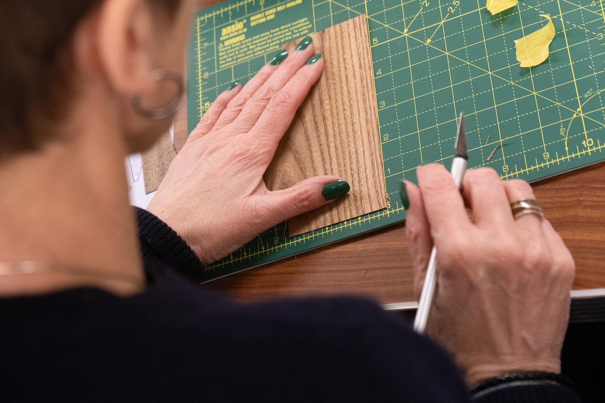 A person with green nail varnish uses a craft knife to cut a piece of wood-look material on a green cutting mat, holding it steady with their other hand.