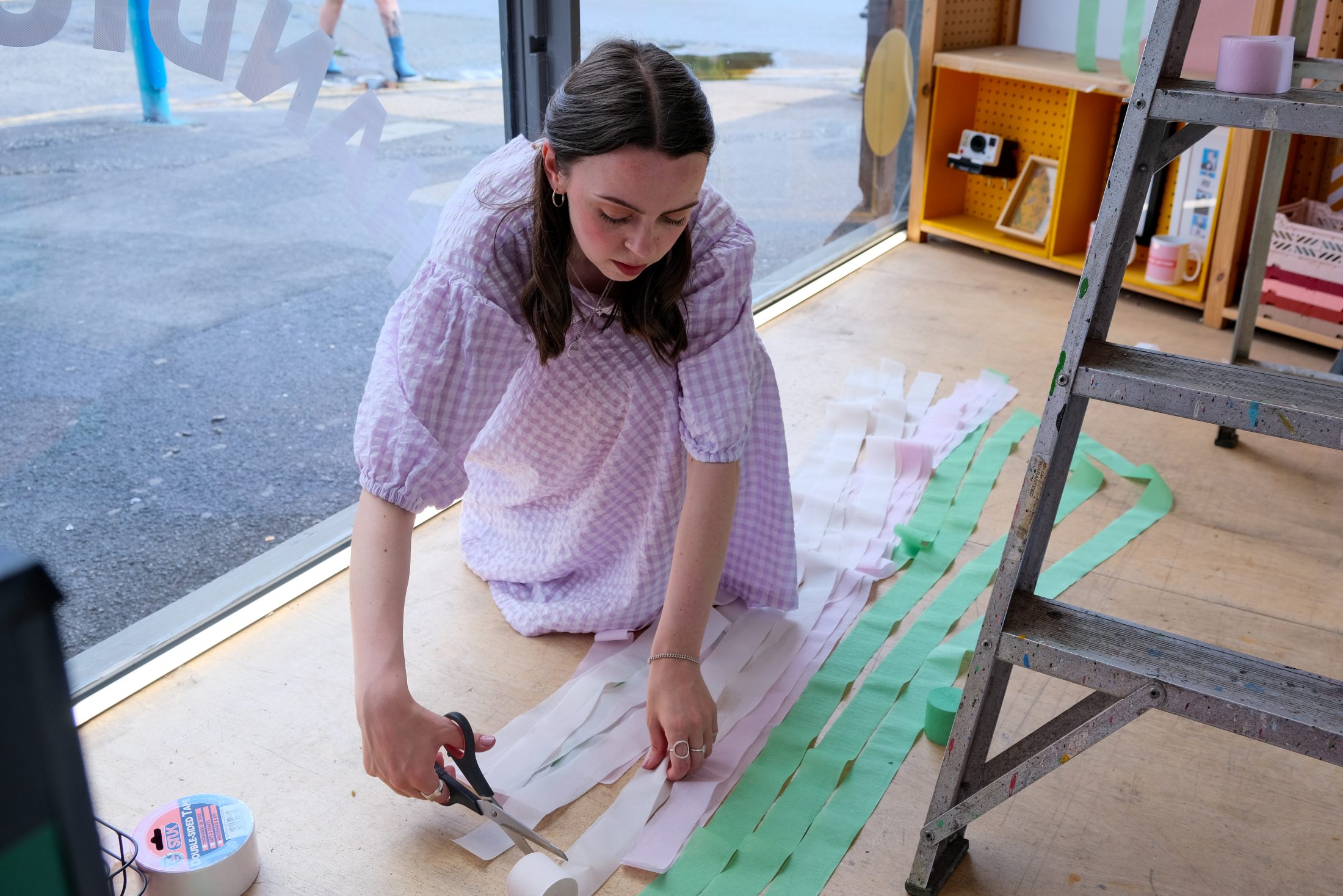 A young woman in a light purple dress kneels on the floor by a window, cutting strips of white and green paper with scissors. Various supplies and a stepladder are nearby.