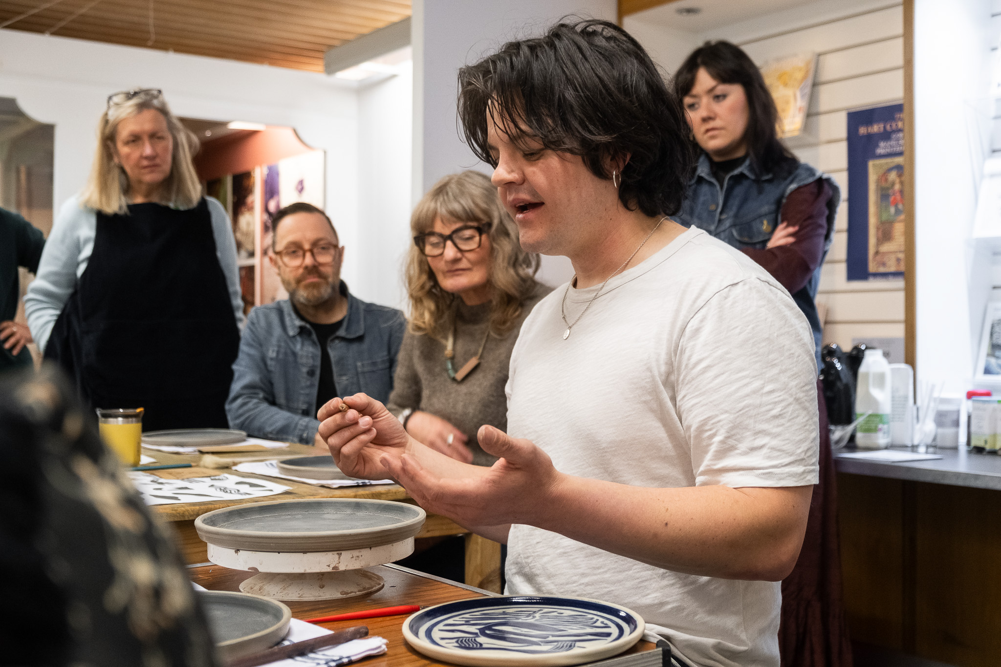 A young man in a white shirt explains pottery techniques to a small group of attentive adults seated around a table, with pottery tools and painted plates in front of him.