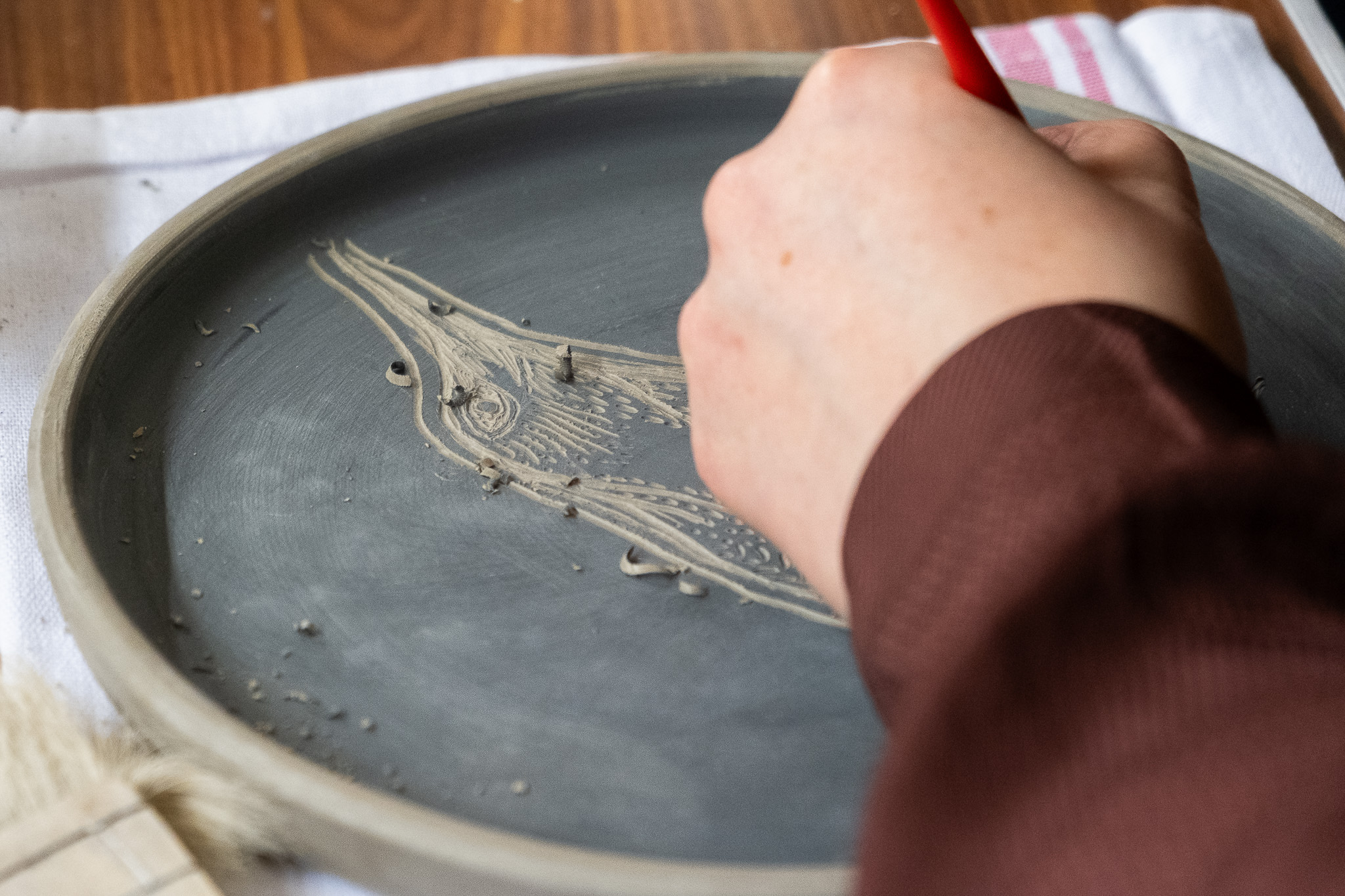 A person uses a pointed tool to carve the outline of a bird onto a round, grey clay plate resting on a table.