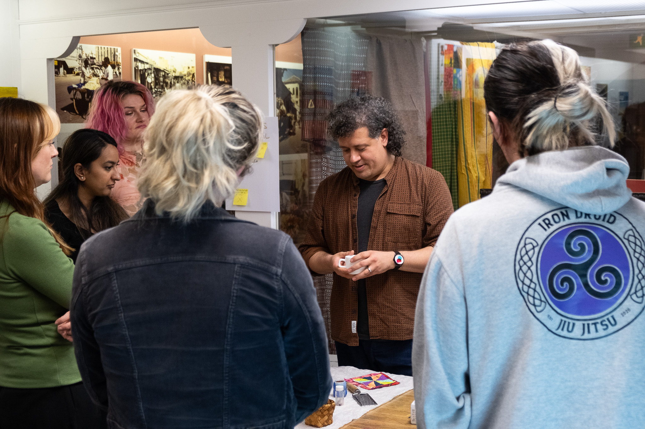 A group of people stand around a table watching a man demonstrate or explain something with his hands; various colourful items are on the table, and the setting appears informal and collaborative.