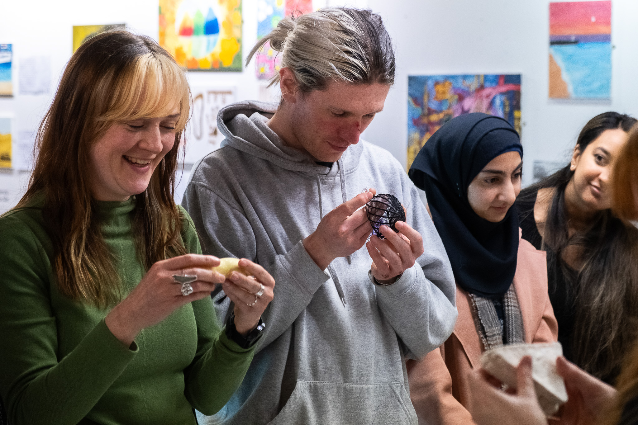 A group of four people, two women smiling and examining objects in their hands, stand together indoors. Colourful artwork is displayed on the wall behind them.