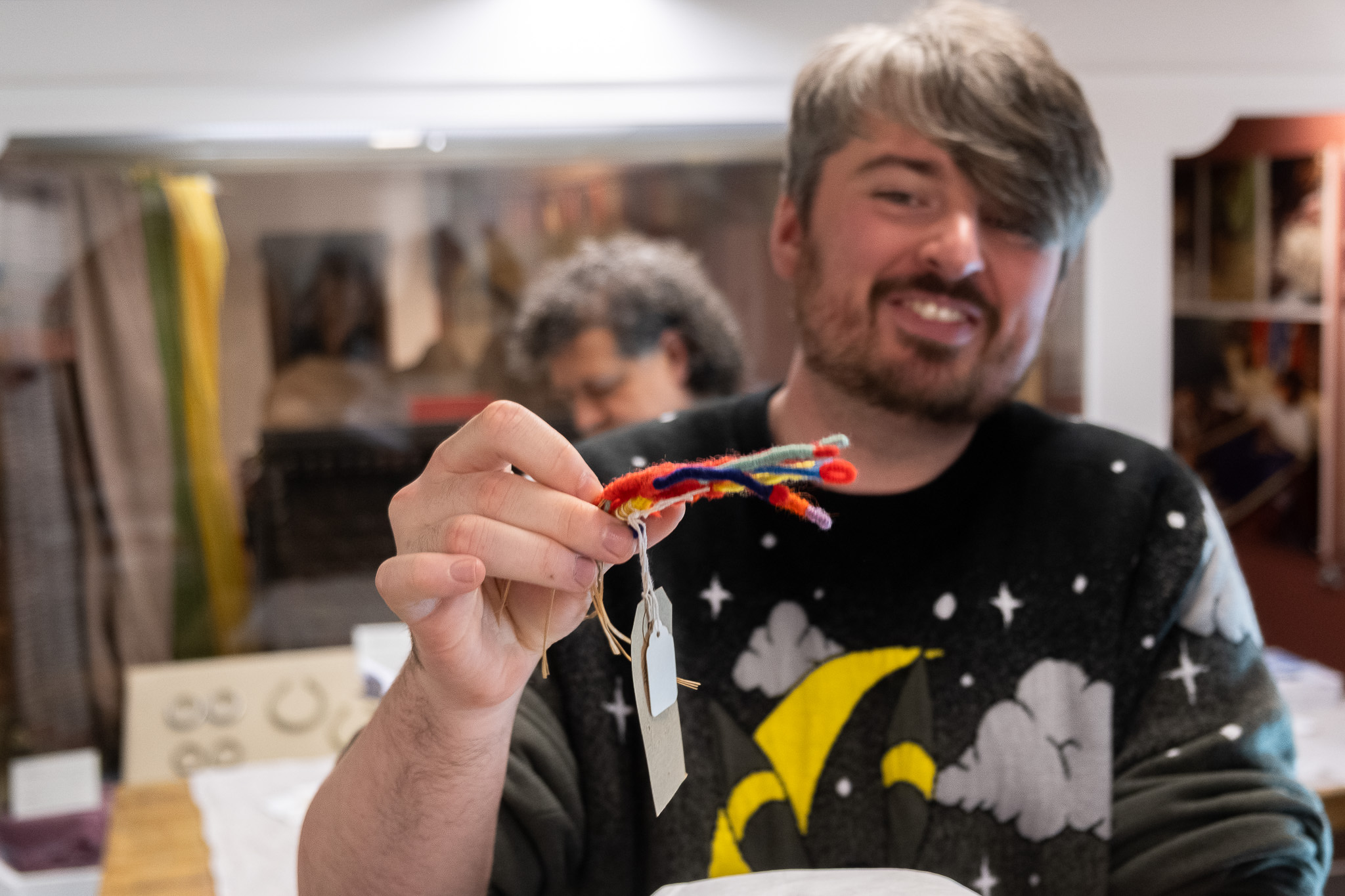 A smiling person with grey hair holds up a small, colourful object made of pipe cleaners and tags in a room with display cases and jewellery in the background.