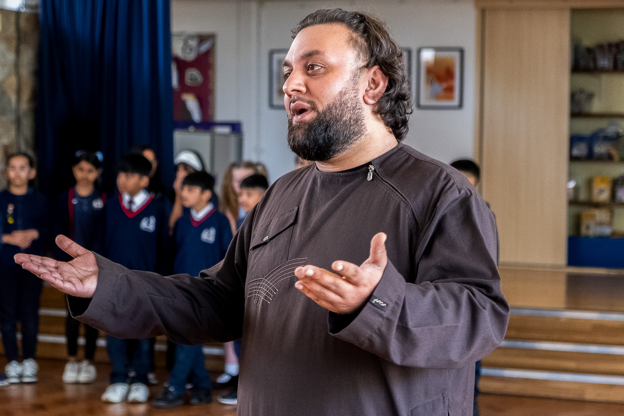 A man with a beard gestures whilst speaking in a classroom setting, with a group of children standing in the background. The children are wearing uniforms and the room has shelves and framed pictures on the walls.