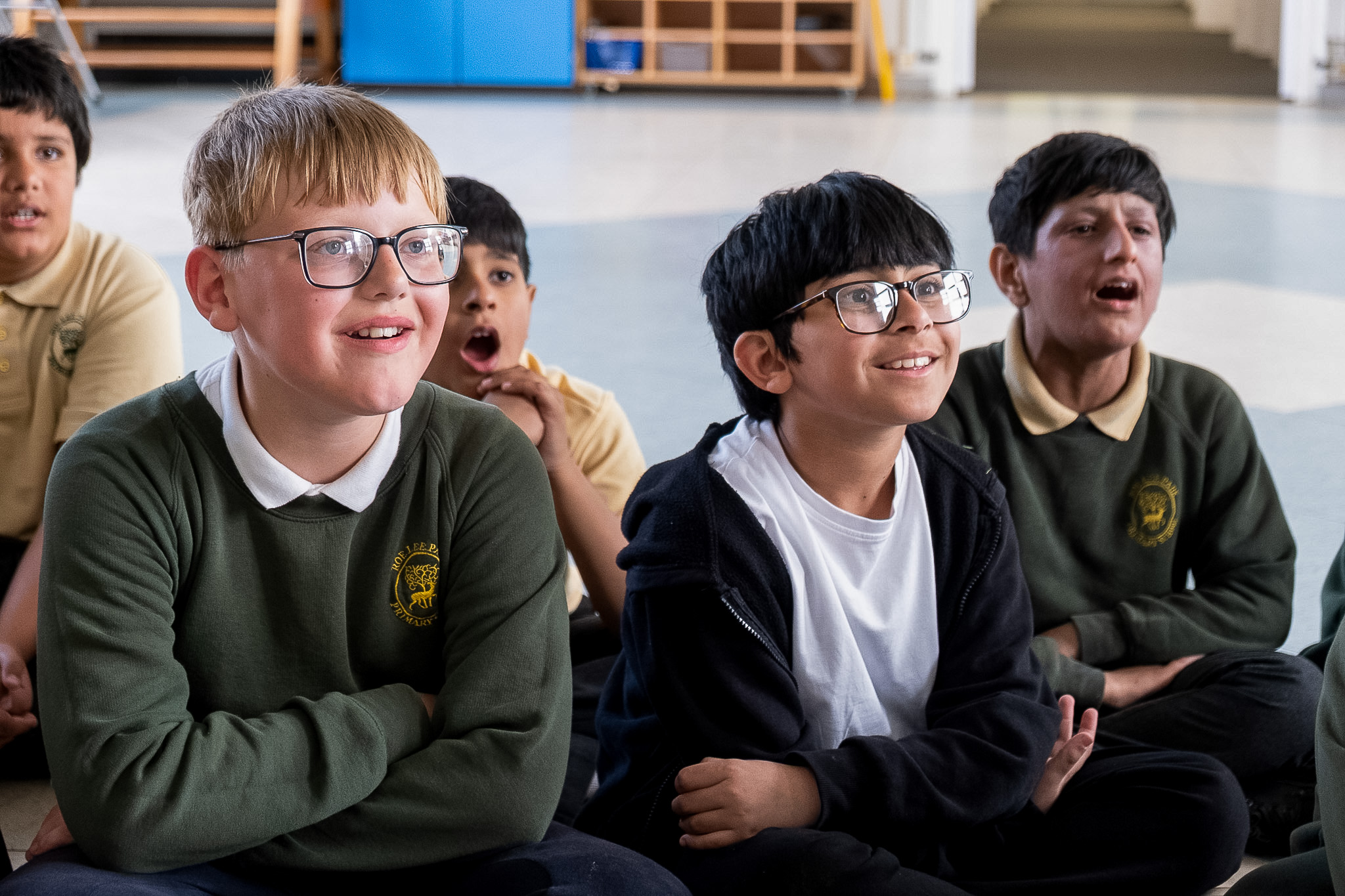 A group of school children sit on the floor indoors, smiling and looking engaged. They are wearing green uniforms and glasses, appearing attentive and happy.
