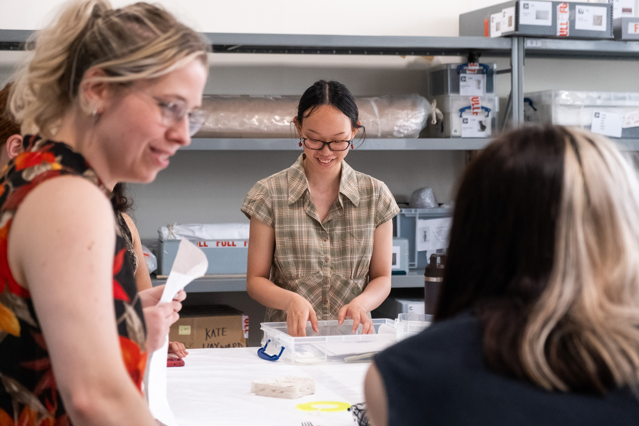 A group gathers around a table working on a project; one person stands smiling, holding an artwork in a clear plastic box, while others sit or stand nearby. Shelves of boxes line the background.