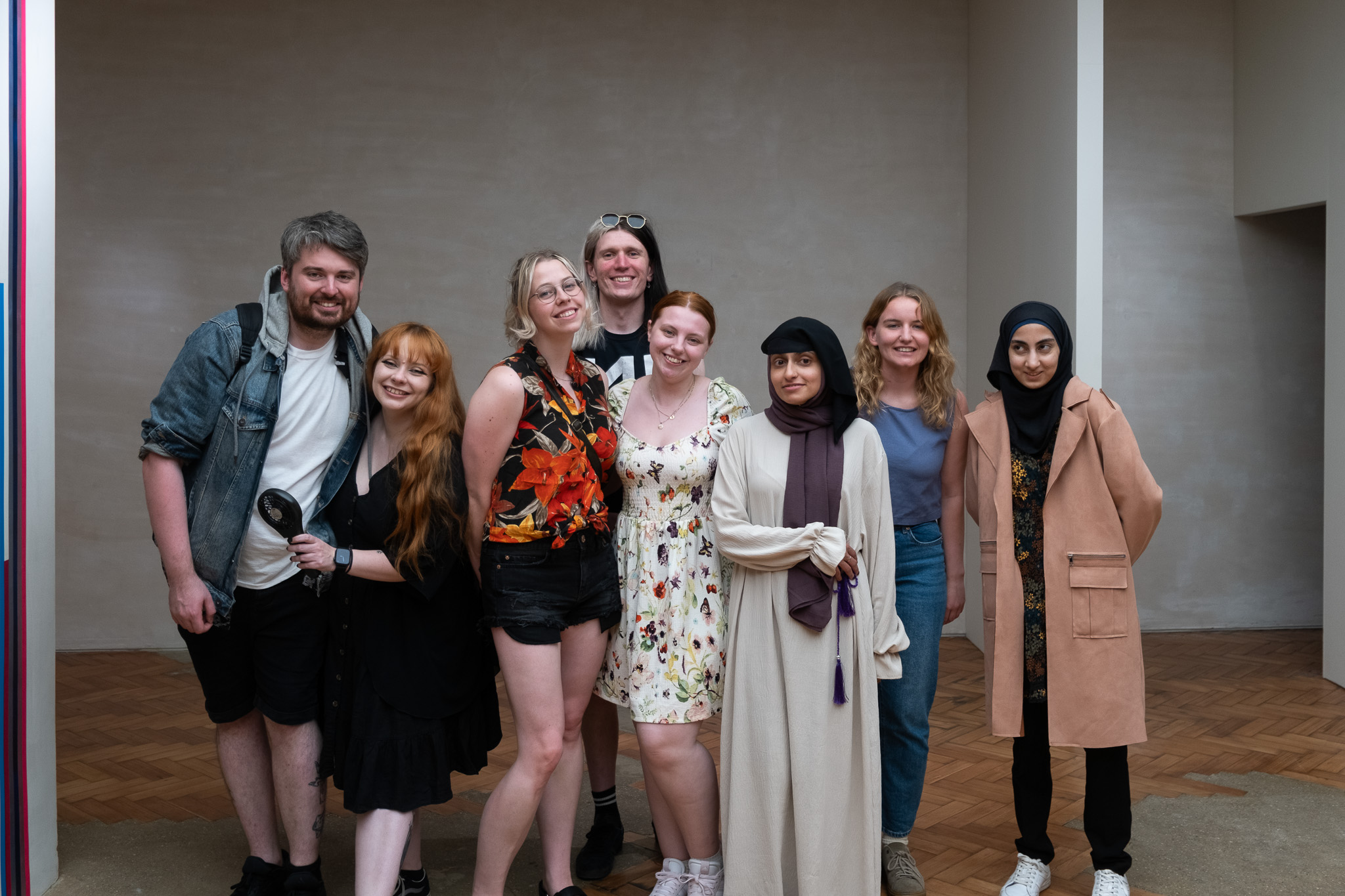 A group of eight young adults posing and smiling together indoors, dressed in casual and summer clothes, standing on a wooden floor with a plain beige wall in the background.