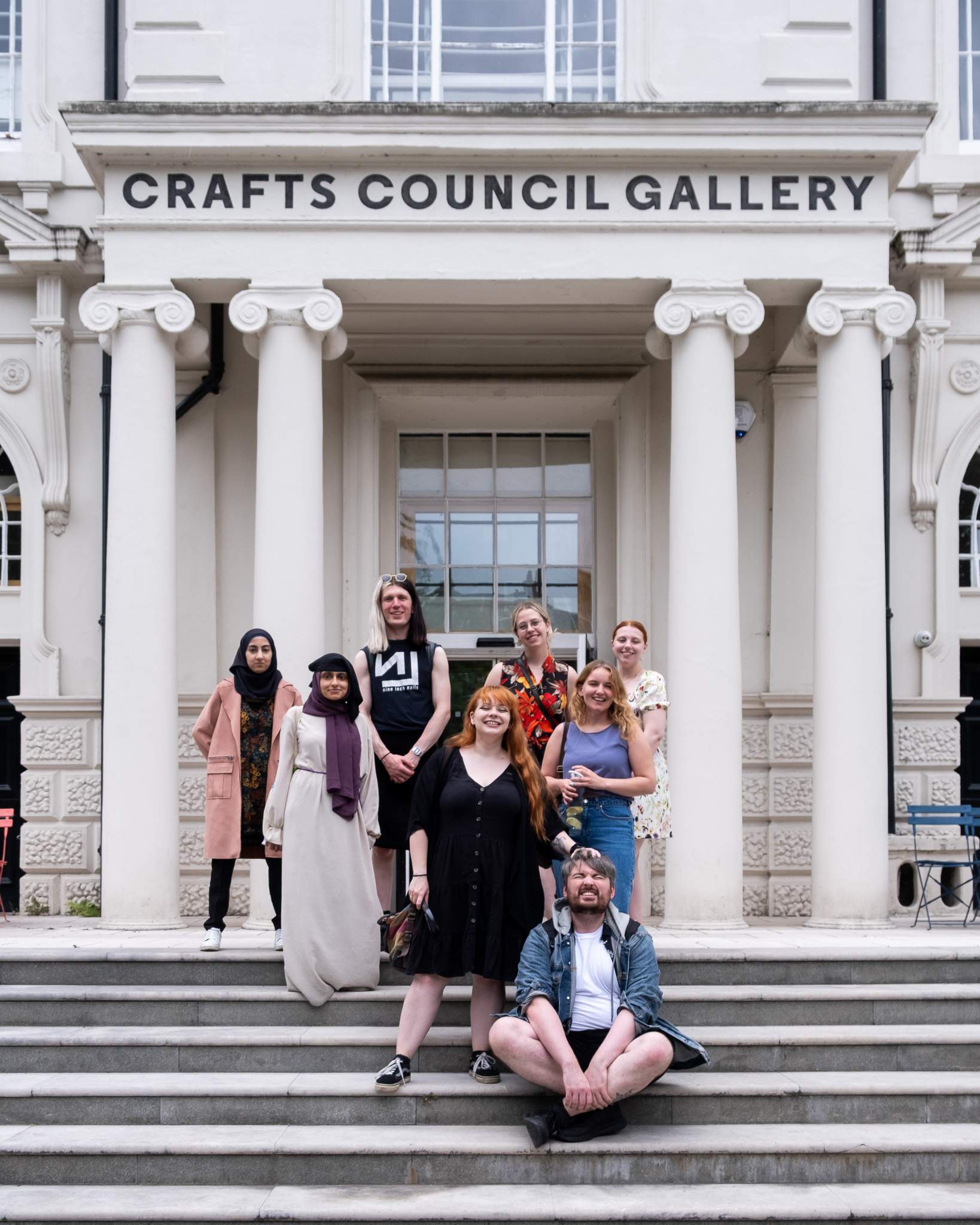 Eight people pose on the steps outside the Crafts Council Gallery building, which has tall white columns and a sign above the entrance. The group appears diverse, with varied ages, styles, and expressions.