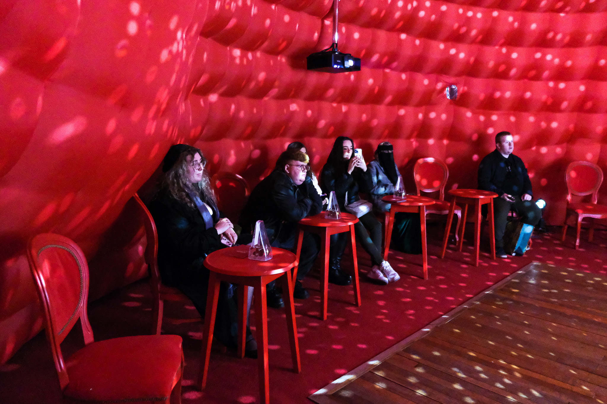 Five people sit at small red tables inside a room with padded red walls and polka-dot lighting effects. The atmosphere is dim, and everyone appears to be watching something ahead.