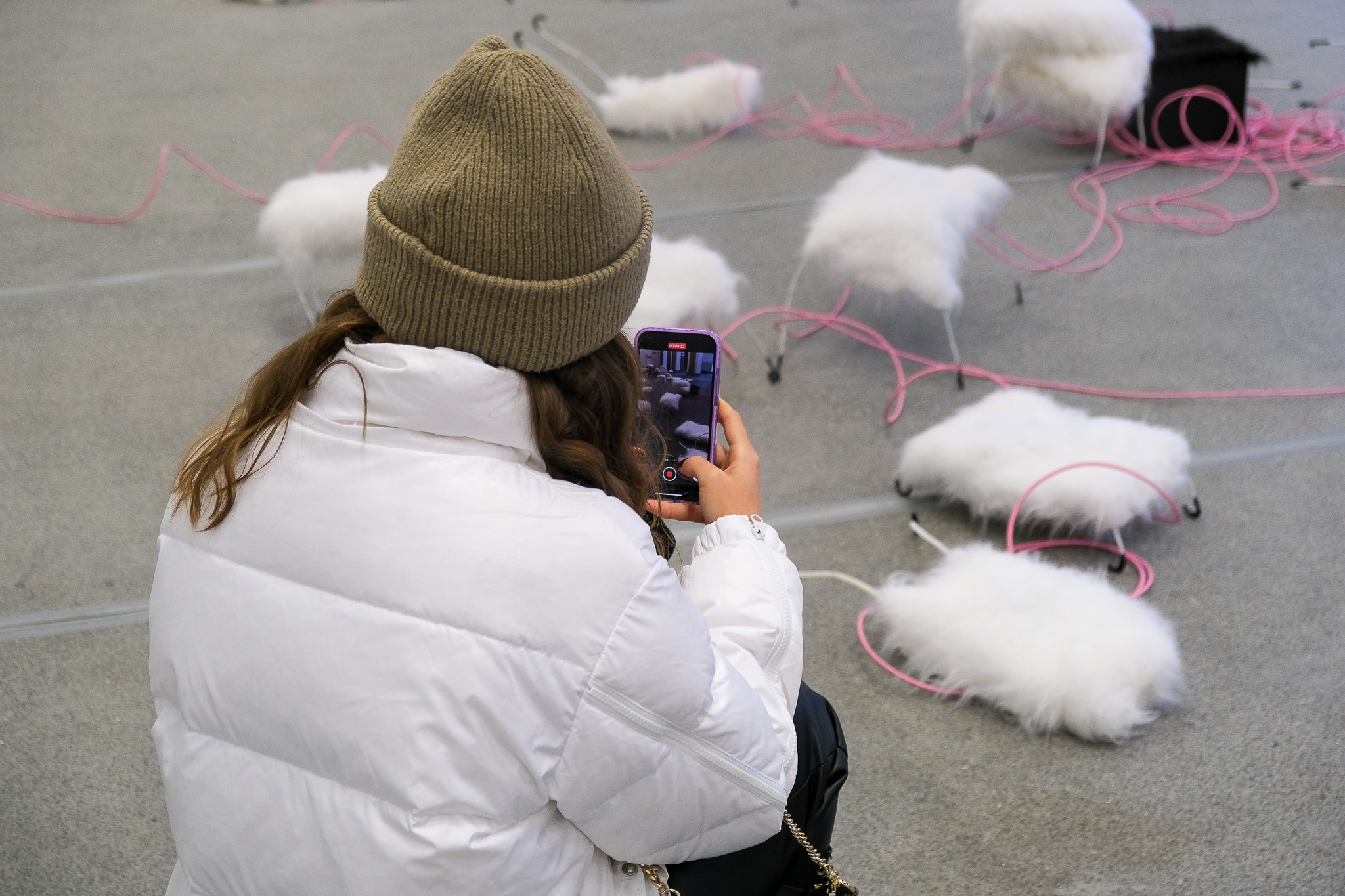 A person in a beige beanie and white puffy jacket kneels and takes a photo with a phone of several fluffy white objects on the floor, connected by pink cables.
