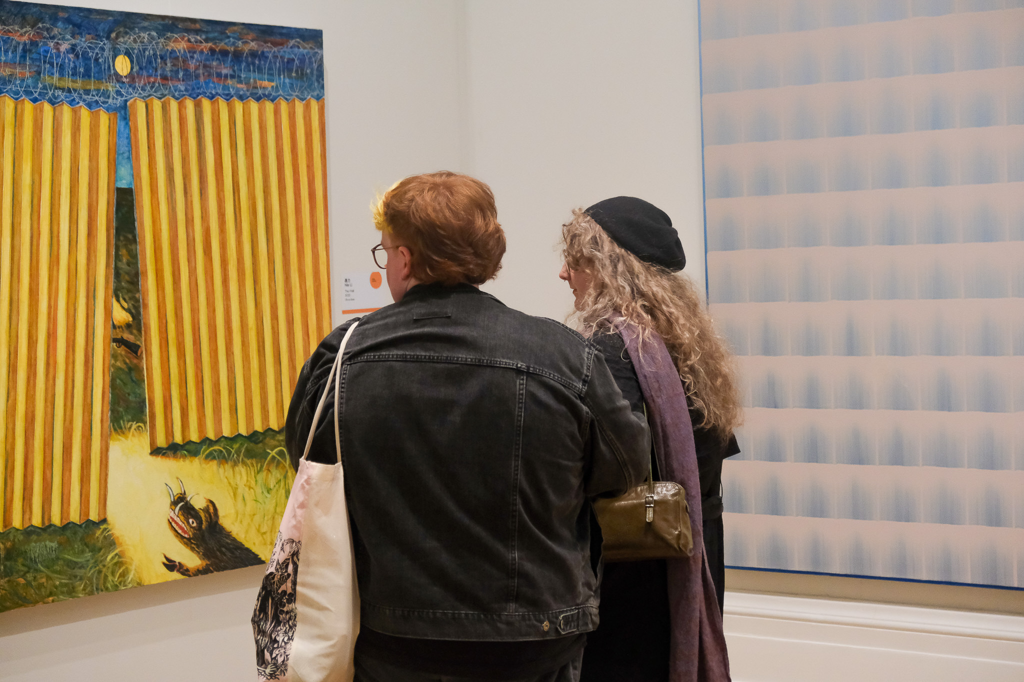 Two people stand closely together in front of colourful framed artwork at a gallery, their backs to the camera as they observe the paintings. One has curly hair and wears a beret; the other wears glasses and a denim jacket.