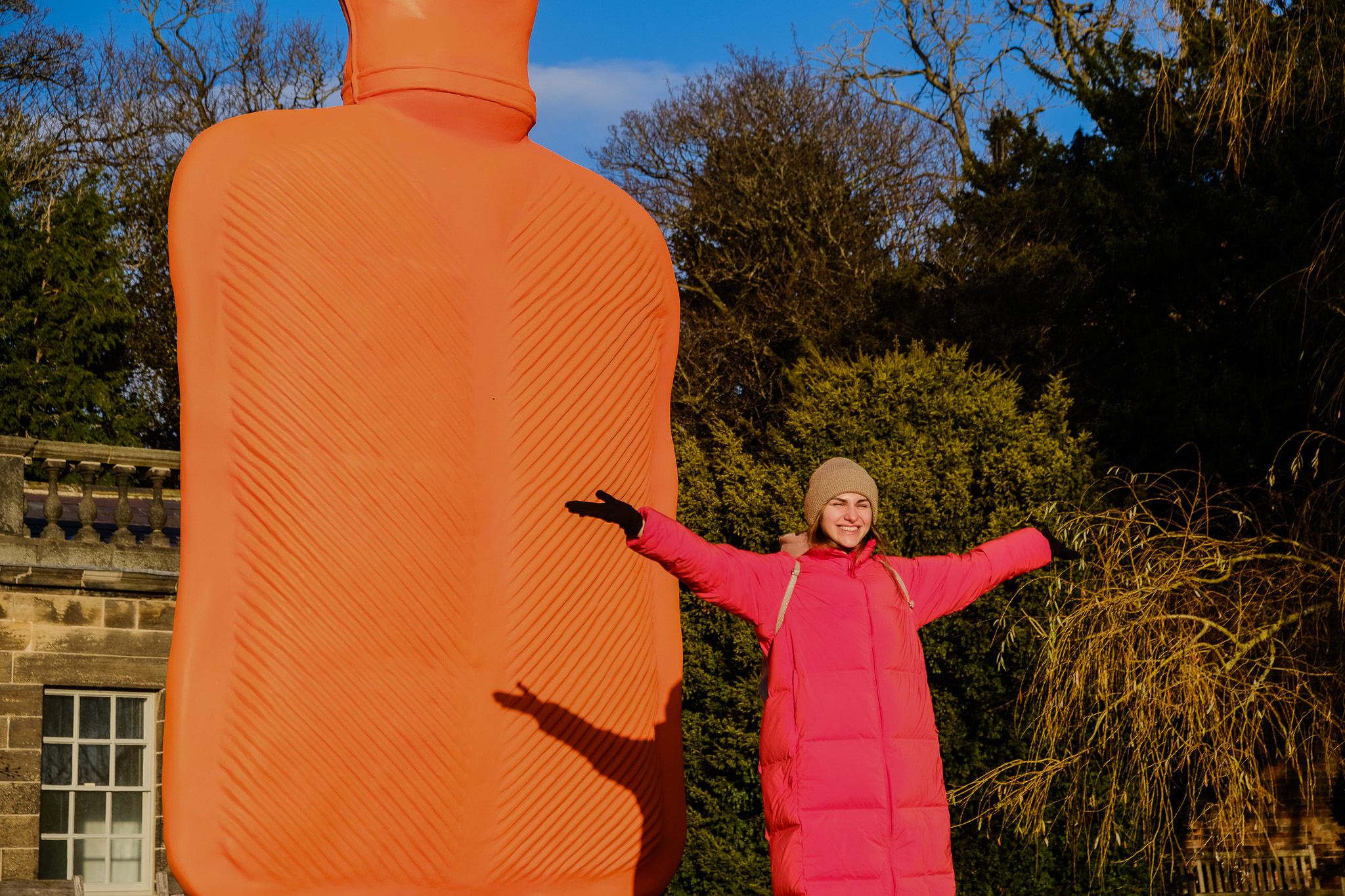 A person in a long pink coat and beige hat stands outdoors with arms outstretched, smiling next to a large orange hot water bottle sculpture on a sunny day with trees and a building in the background.