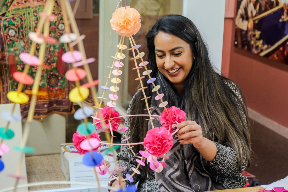A woman with long dark hair smiles as she crafts a colourful hanging decoration with paper flowers and circular cut-outs in a bright, indoor setting.