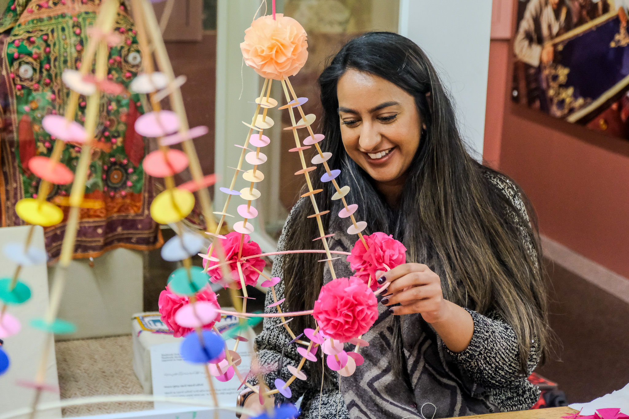 A woman with long dark hair smiles as she crafts a colourful hanging decoration with paper flowers and circular cut-outs in a bright, indoor setting.