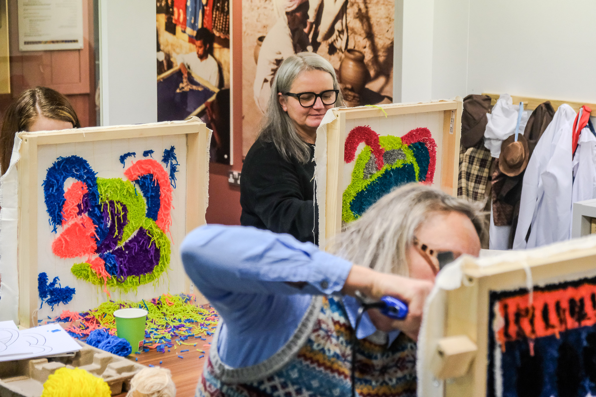 Two people work on colourful tufted yarn art pieces in wooden frames at a craft workshop. Yarn scraps are scattered on the table. Photos and clothing hang on the walls in the background.