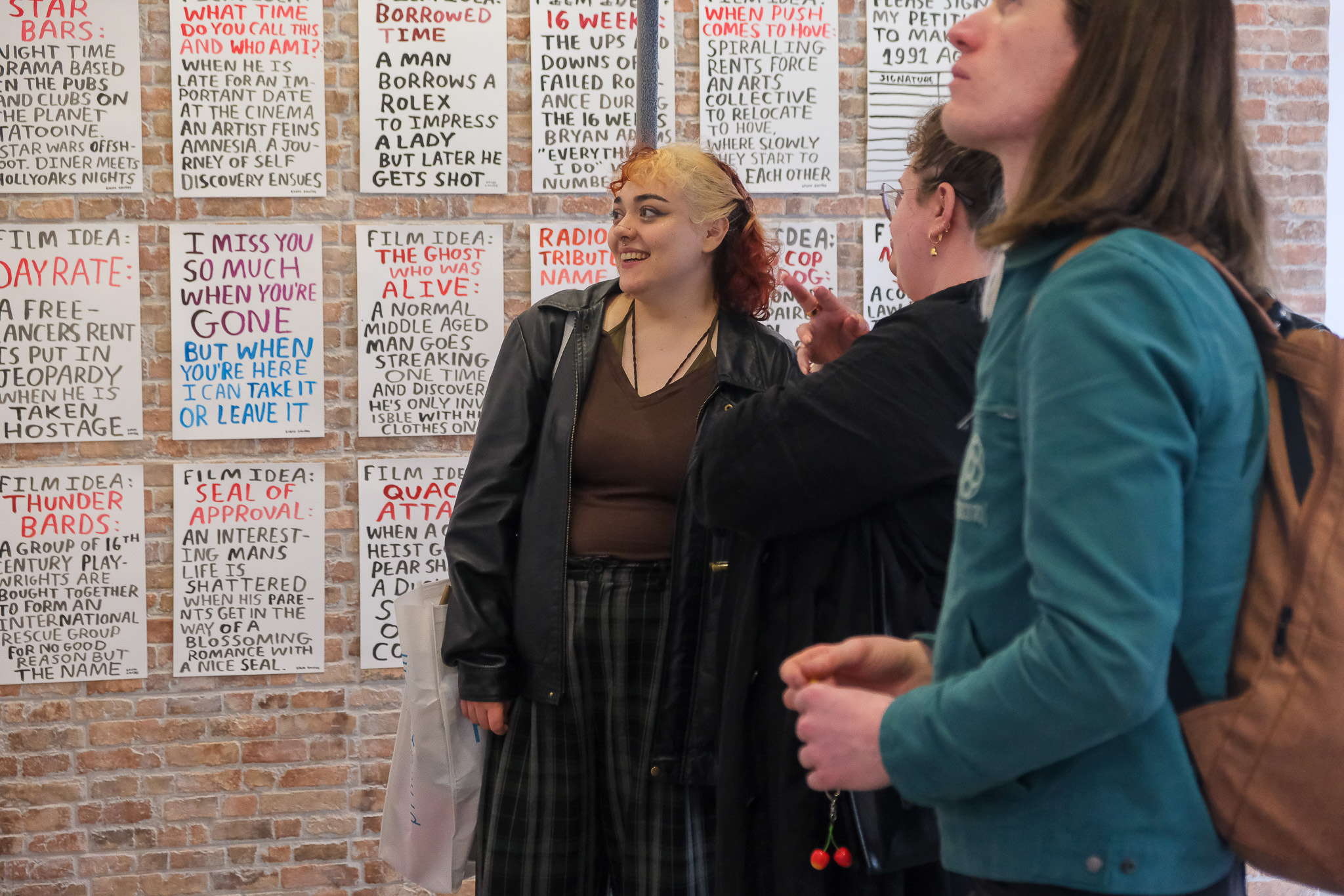 Three people stand and look at a wall covered with white posters featuring handwritten red and black text. One person smiles, and another holds cherry-shaped earrings. The background is a brick wall.