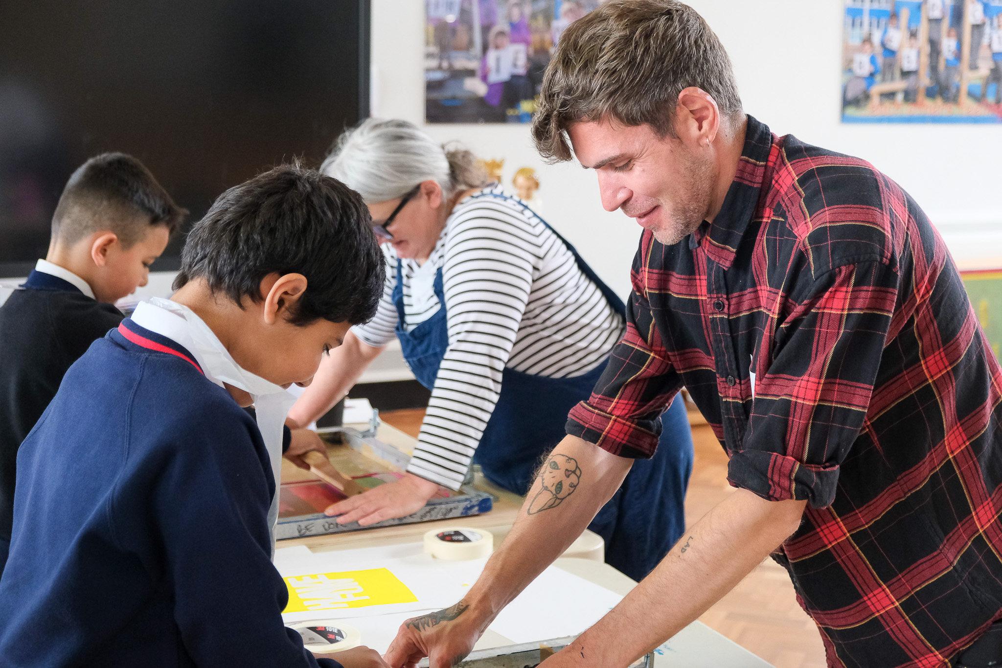 A man in a red check shirt helps children with screen printing at a table, while another adult works alongside them in a classroom setting.