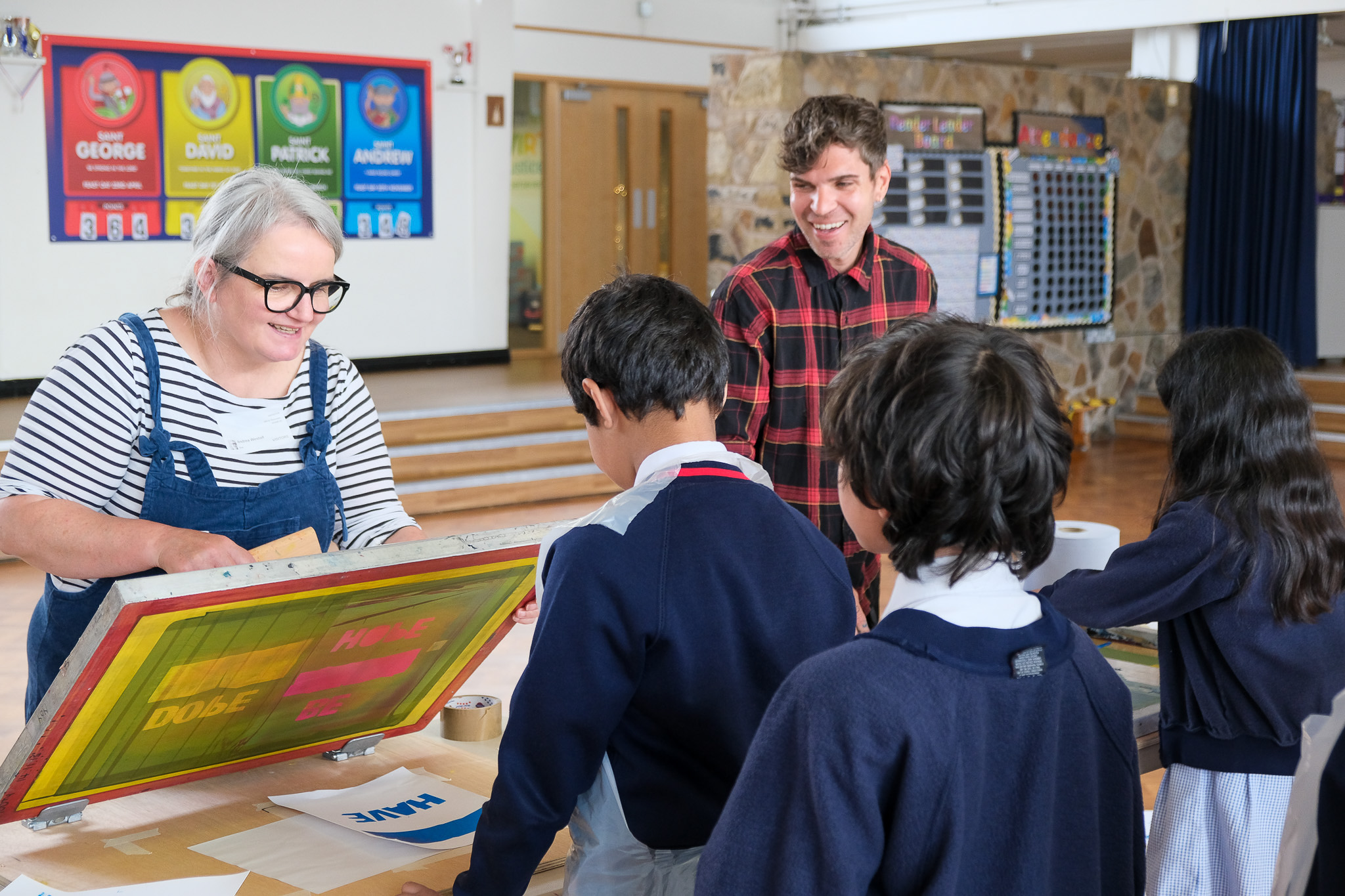 A woman demonstrates screen printing to a group of schoolchildren while a man observes and smiles in a classroom. The children wear uniforms and posters with names are visible on the wall in the background.