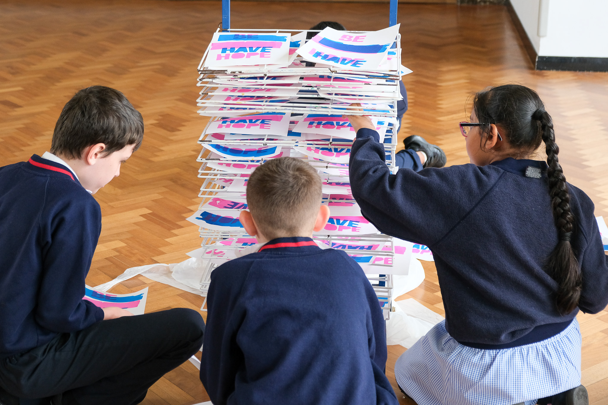 Three children sit on a wooden floor, placing colourful printed sheets onto a drying rack during an art activity.