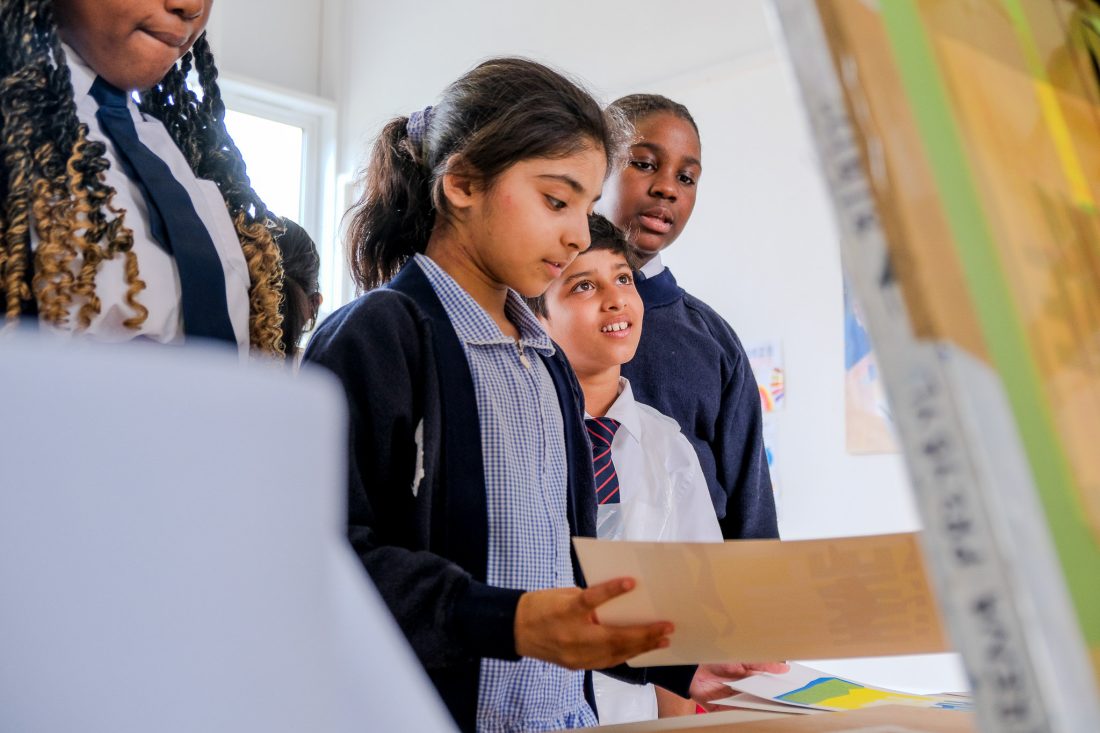 Four schoolchildren wearing uniforms stand together indoors, looking at artwork or papers with interest. One girl in the foreground holds a sheet, while the others observe attentively. The setting appears to be a classroom or art space.
