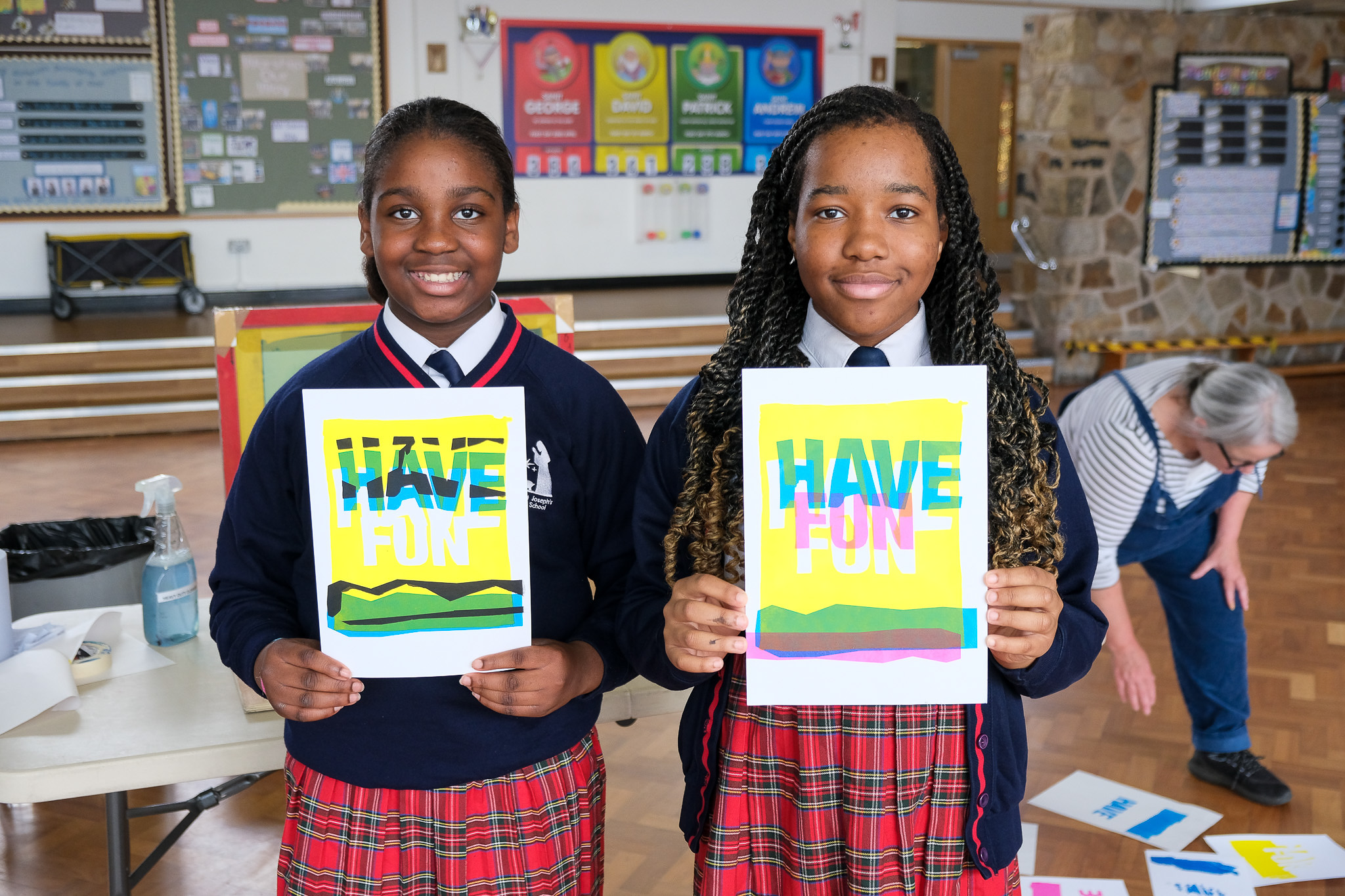 Two girls in school uniforms stand indoors, holding colourful posters that say HAVE FUN. They are smiling at the camera, and in the background, a teacher is arranging similar posters on the floor.
