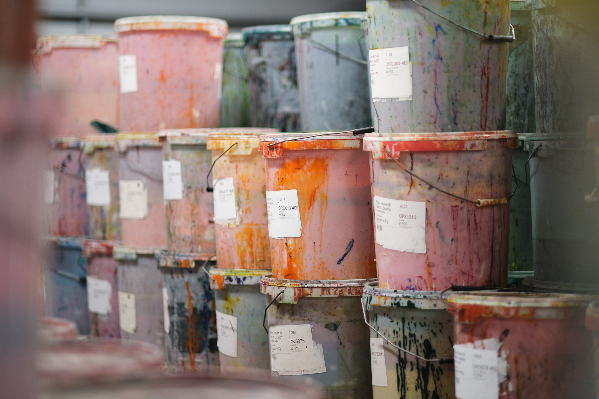 Stacks of large plastic buckets covered in dried, colourful paint drips and smudges, each labelled with white paper. The buckets are arranged in rows, creating a vibrant, messy pattern.
