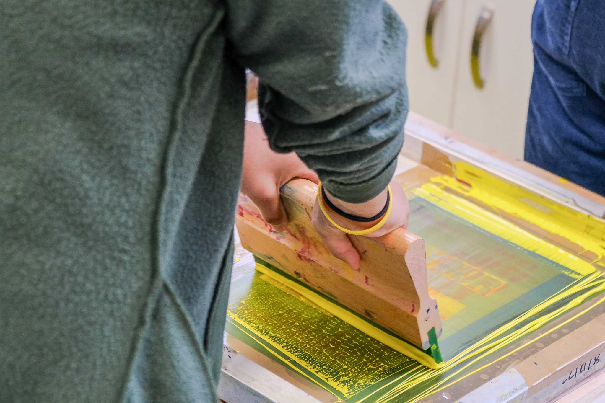 A person uses a wooden squeegee to spread yellow ink over a screen printing frame, creating a design on paper or fabric. The focus is on the hands and tool, with the process in progress.