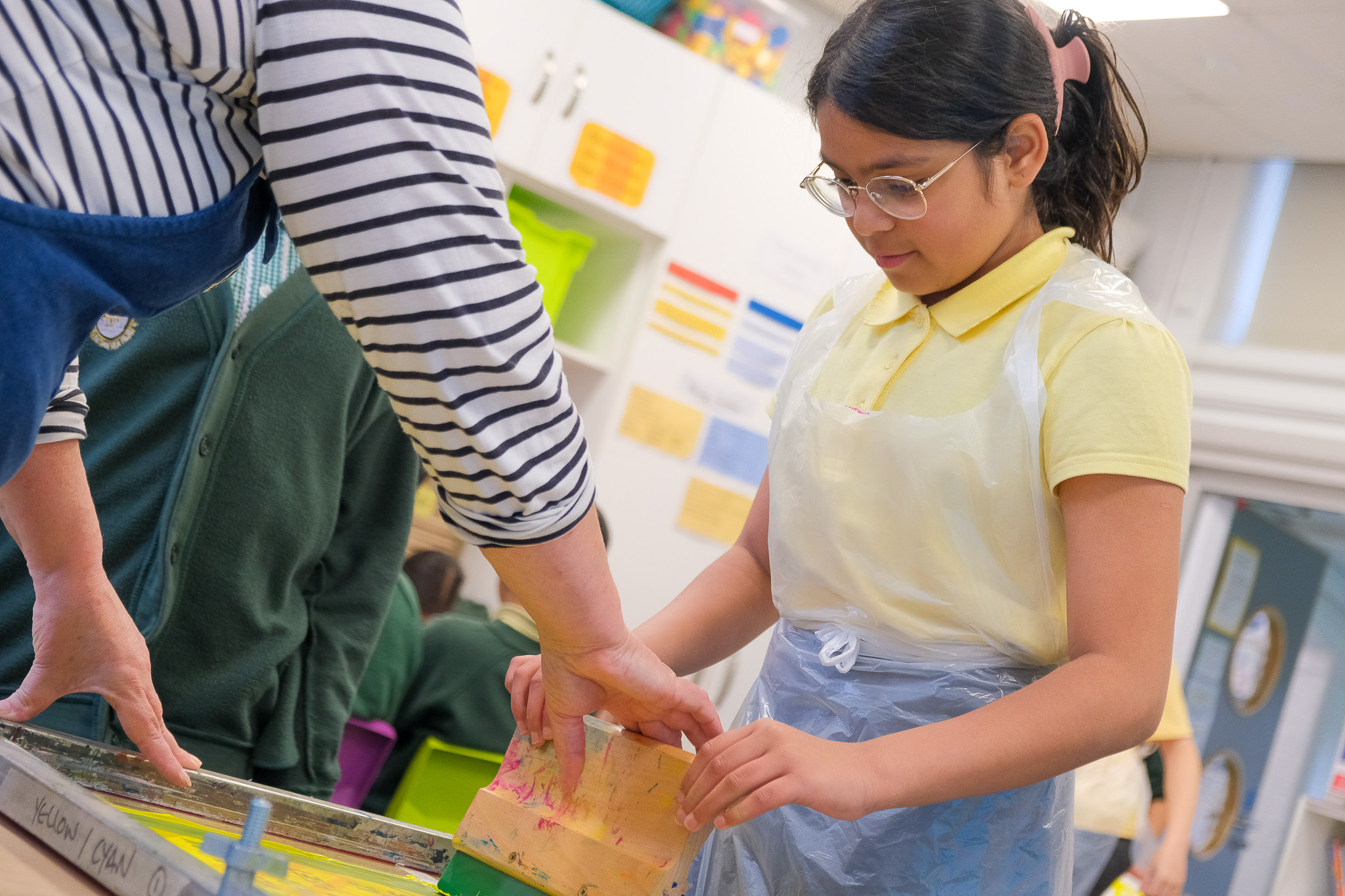 A girl wearing glasses and a yellow shirt, covered with a plastic apron, works on an art project in a classroom, assisted by an adult in a striped shirt. Other pupils are visible in the background.