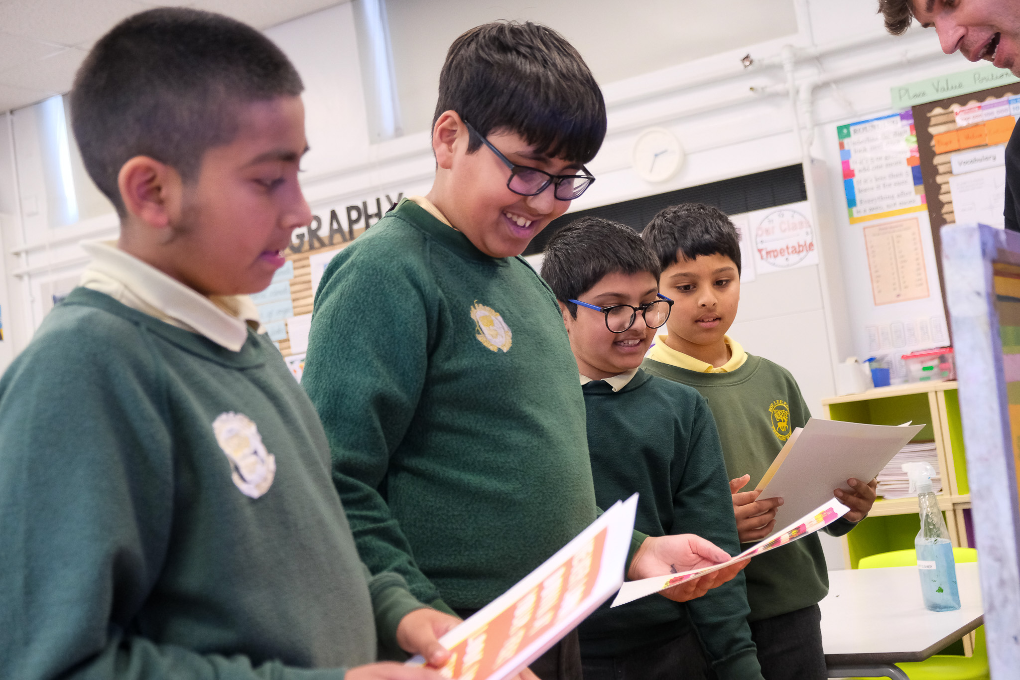 Four boys in school uniforms stand in a classroom, smiling and looking at colourful papers they are holding. Classroom posters and charts are visible on the walls in the background.