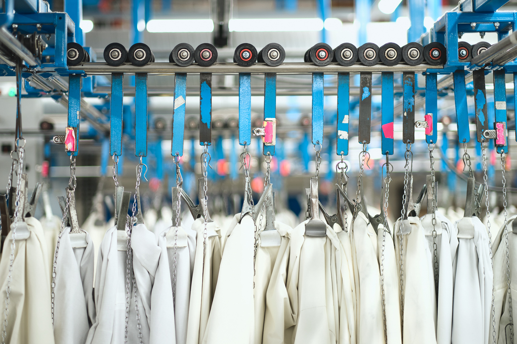 Several white garments hang on metal hangers attached to a conveyor system in a brightly lit industrial laundry or factory setting, with blue rails and wheels above them.