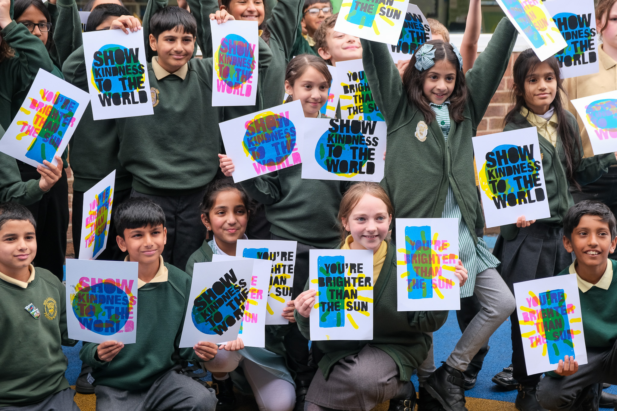 A group of smiling schoolchildren in green uniforms hold up colourful posters with positive messages like “SHOW KINDNESS TO THE WORLD” and “YOU’RE BRIGHTER THAN THE SUN”.
