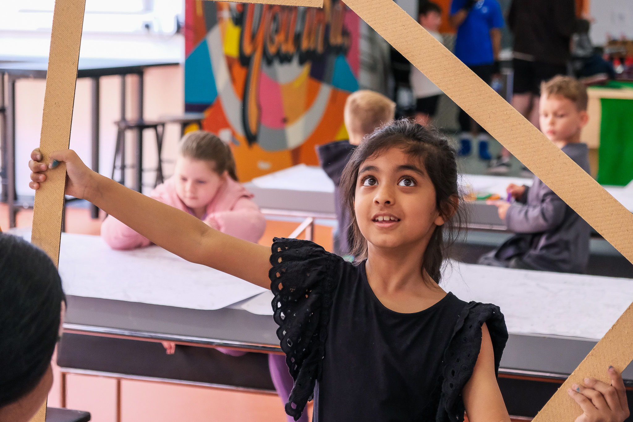A young girl in a black dress holds up a large cardboard frame. Other children are seated and engaged in activities.