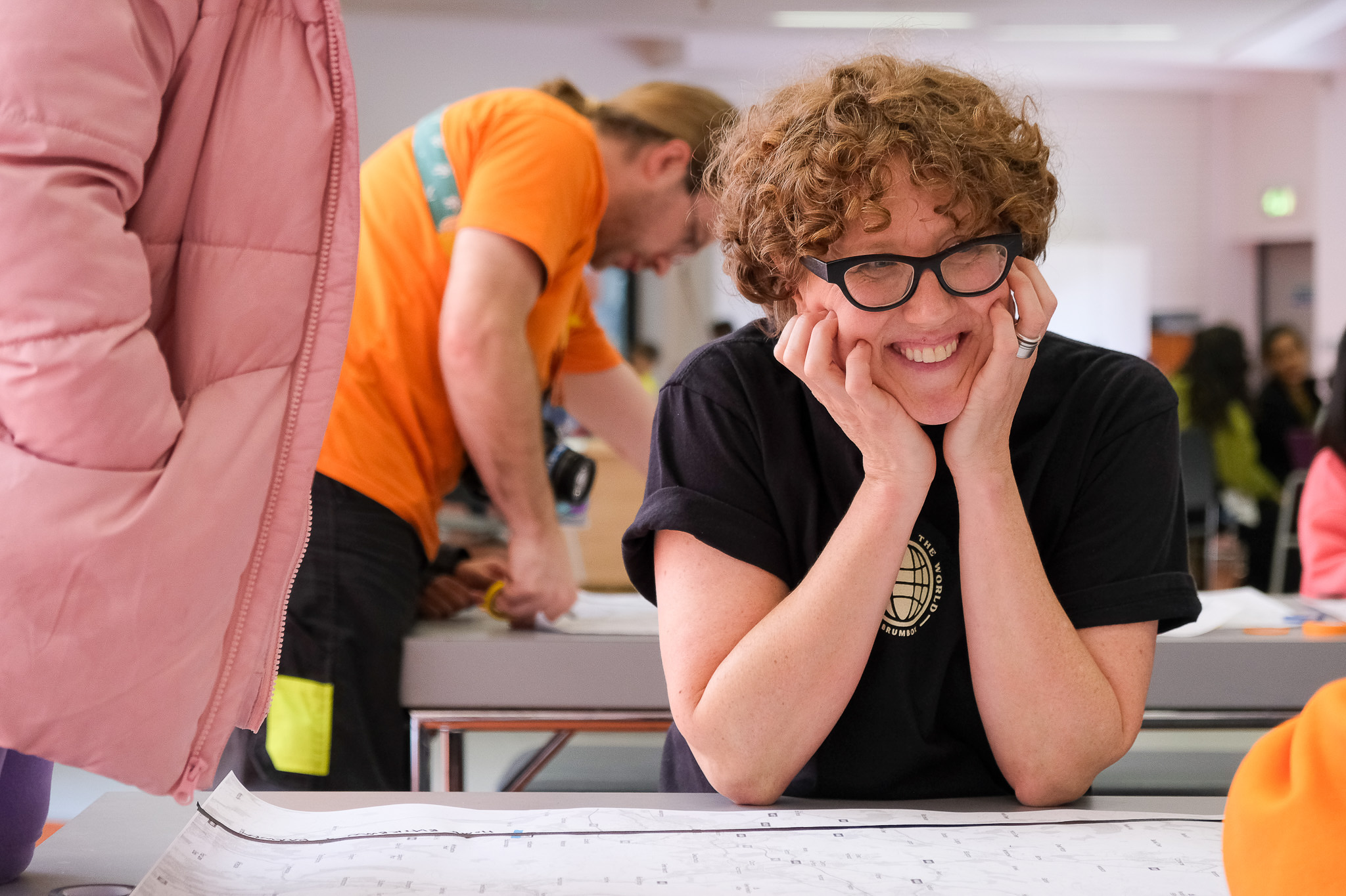 A smiling person with curly hair and glasses leans on a table, looking at someone off-camera. They are indoors, and other people are working in the background.