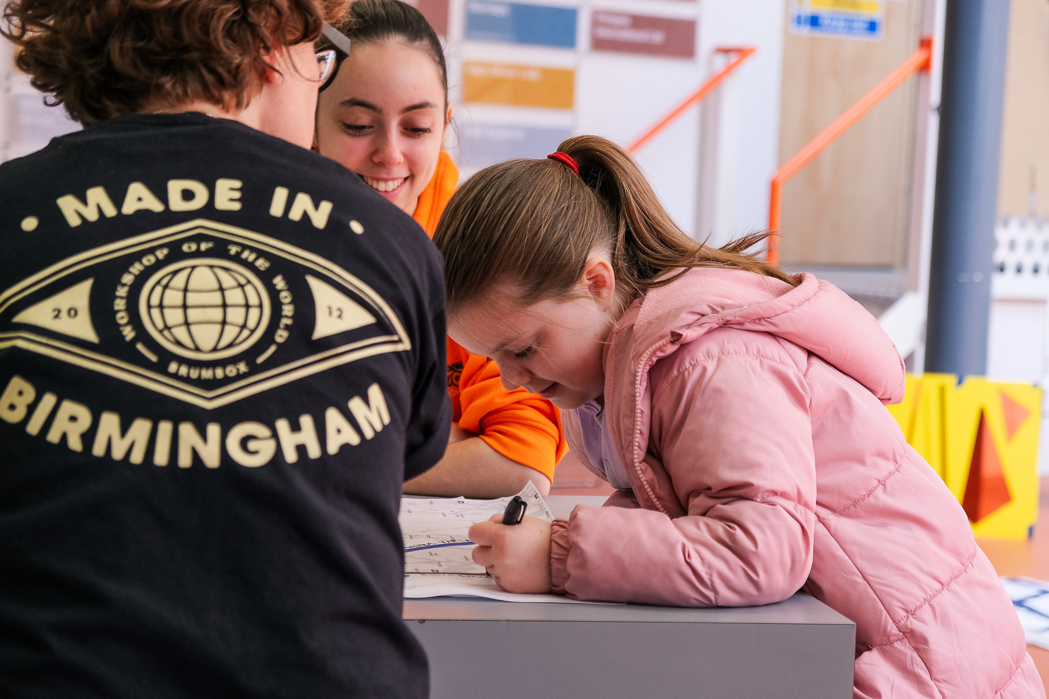 A girl in a pink coat leans over a table, drawing on paper, whilst two adults stand nearby. One adult wears a Made in Birmingham shirt and the other smiles, watching the girl.
