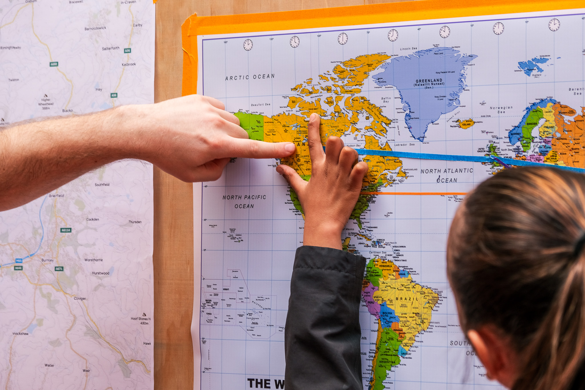 Two people point at a location on a world map, specifically in North America, while another map is partially visible on the left. One person’s arm is extended whilst another uses both hands to touch the map.