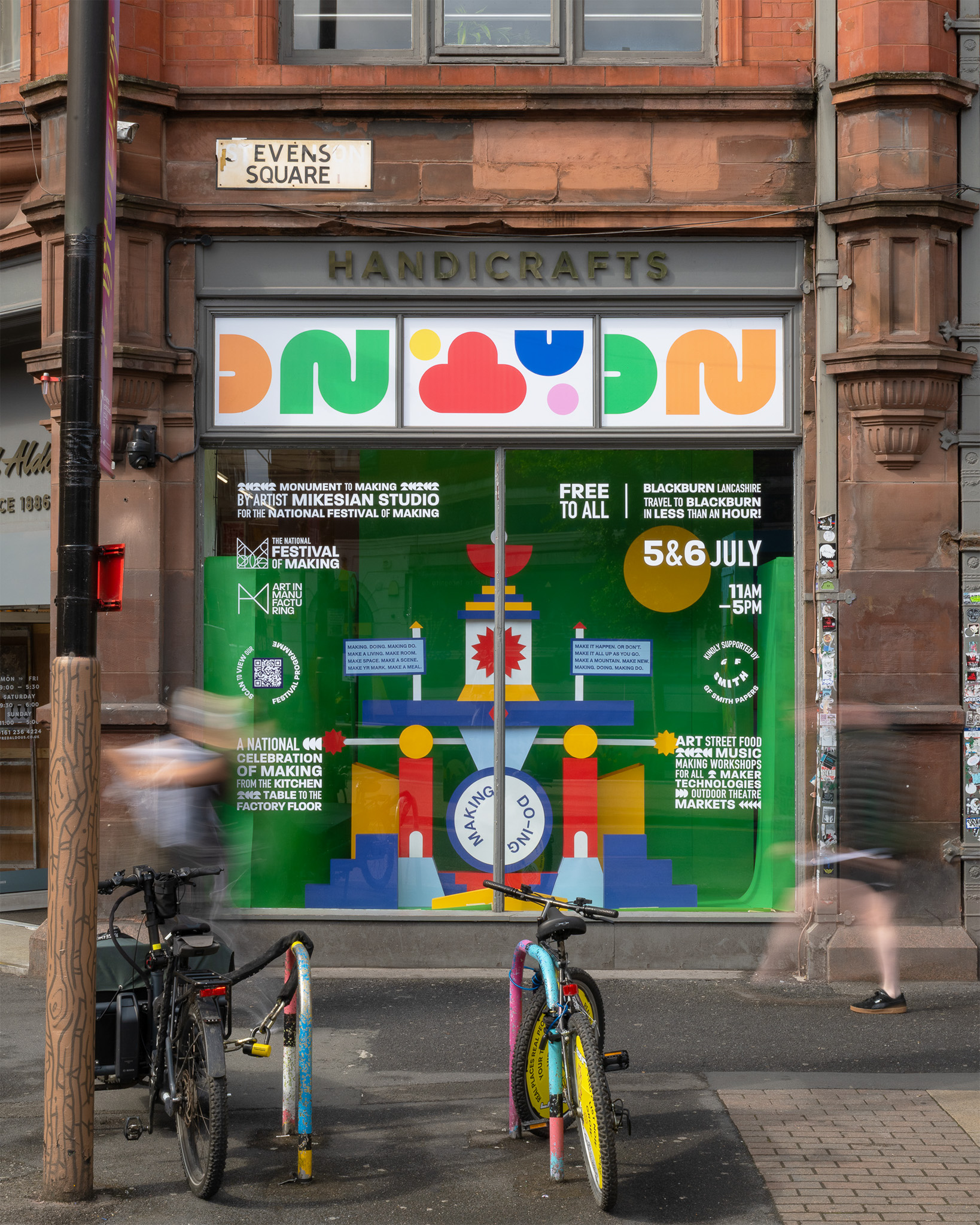 A colourful shopfront window advertises a free festival with bold graphics and event details. Two bicycles are parked outside, and blurred pedestrians walk past the HANDICRAFTS shop sign on a red brick building.