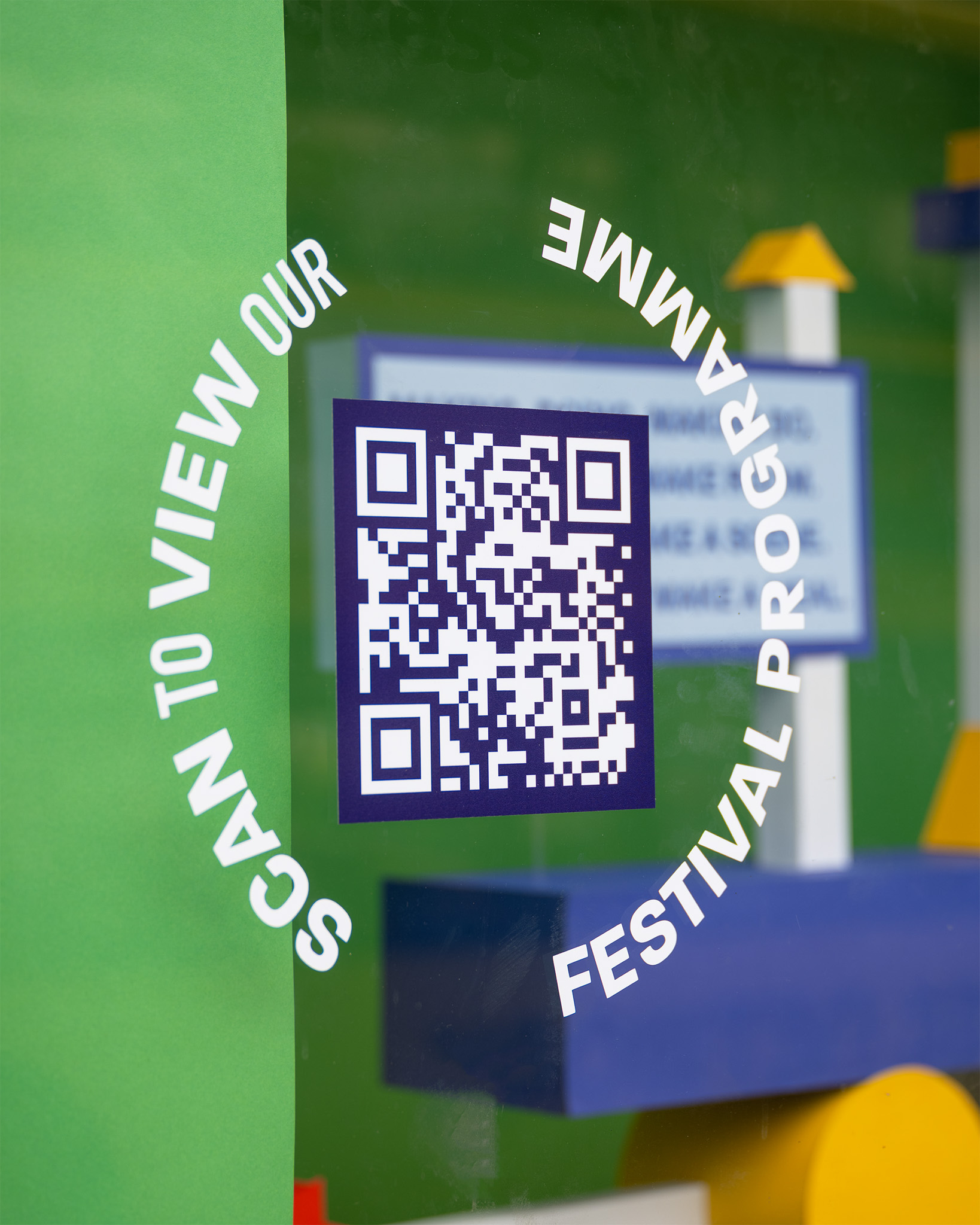 A circular sign with a QR code in the centre reads “SCAN TO VIEW OUR FESTIVAL PROGRAMME,” displayed on a window with colourful abstract shapes in the background.