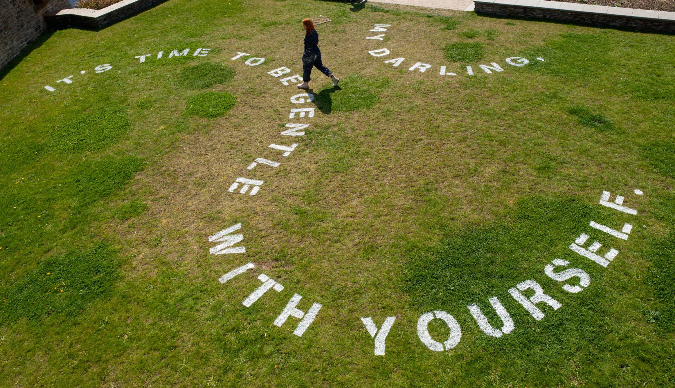 A person walks across a grassy area where large white letters spell out, “It’s time to be gentle with yourself, my darling.” The text forms a spiral pattern on the grass.