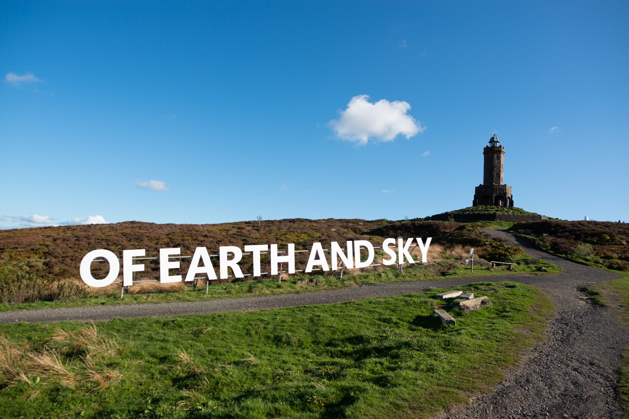 Large white letters spell “OF EARTH AND SKY” on grassy land beside a gravel path. In the background, a stone tower stands atop a hill under a bright blue sky with a few clouds.