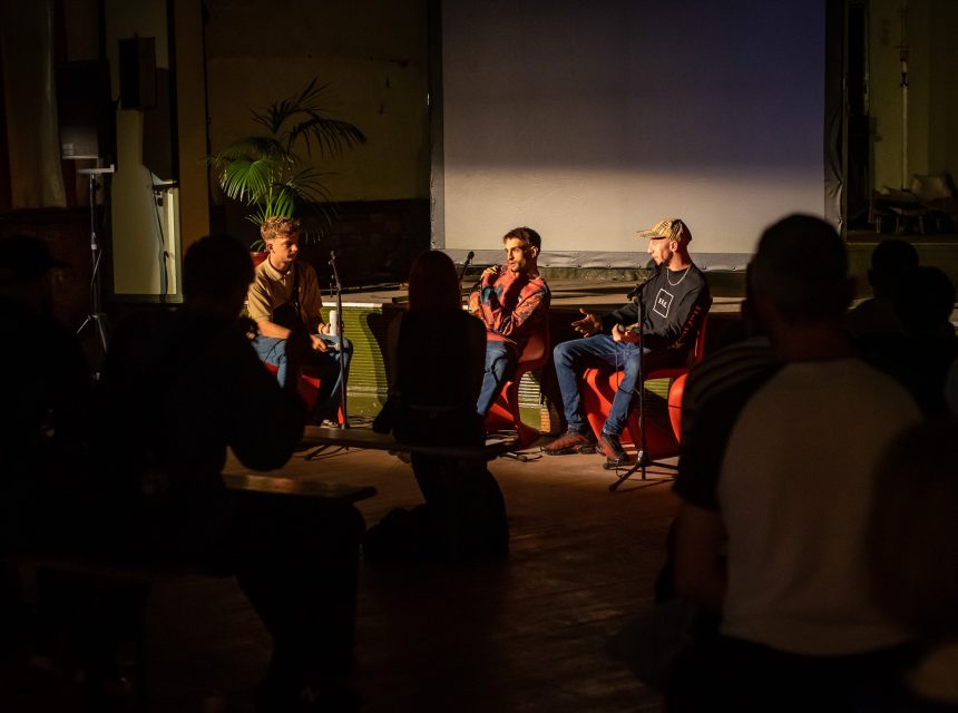 Three people sit on stage having a discussion in front of an audience in a dimly lit room. One person speaks into a microphone whilst the others listen. The audience is seated on benches, and a large screen is behind the speakers.