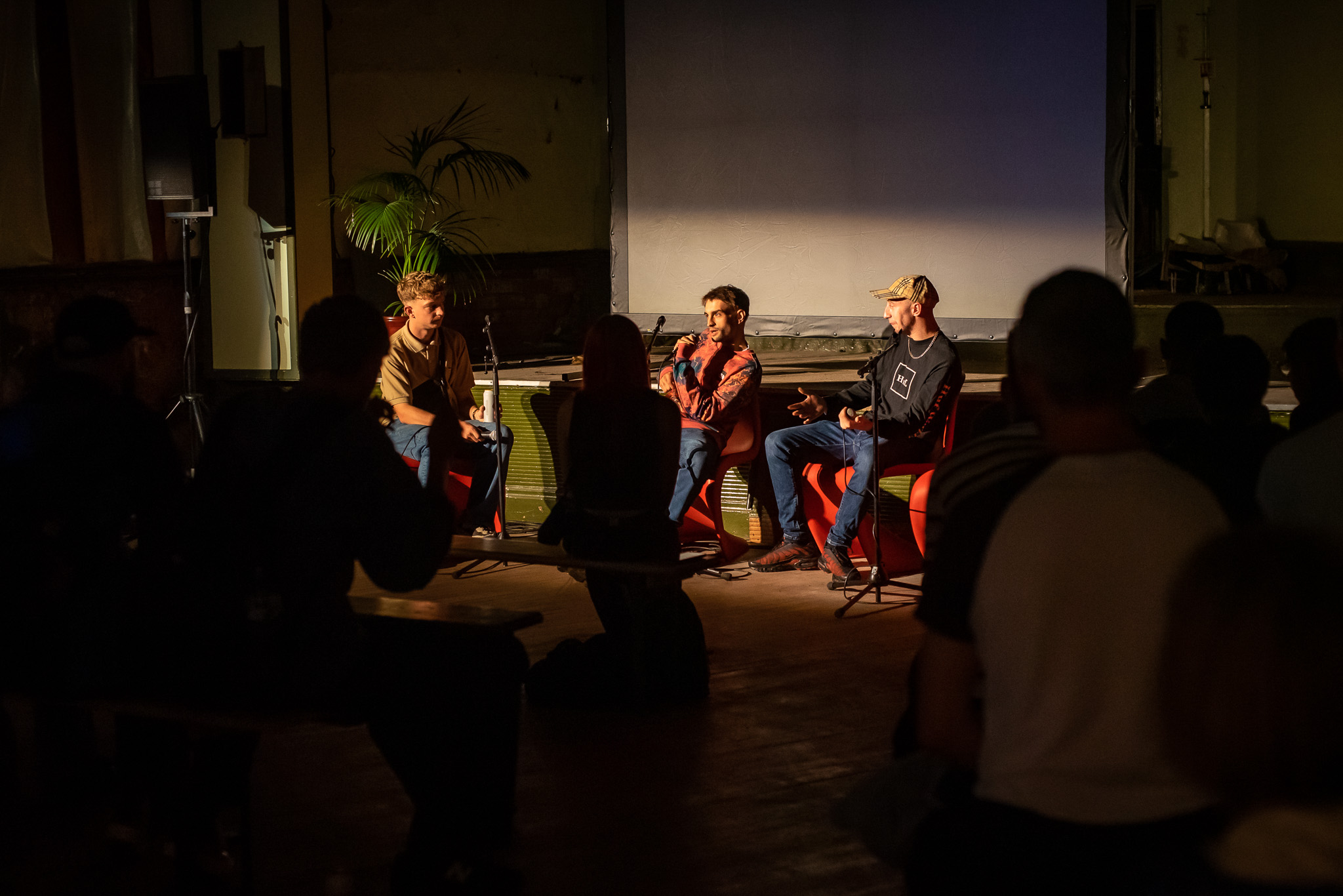 Three people sit on stage having a discussion in front of an audience in a dimly lit room. One person speaks into a microphone whilst the others listen. The audience is seated on benches, and a large screen is behind the speakers.