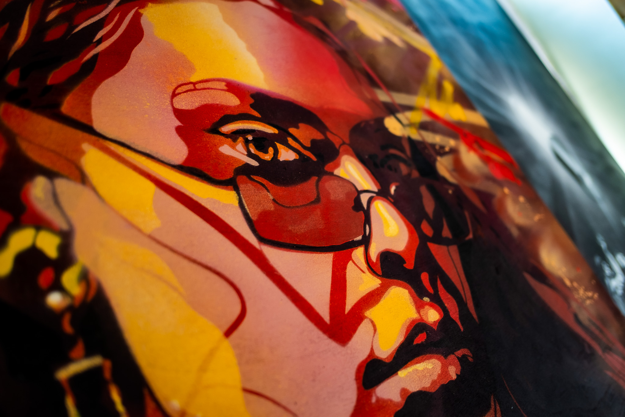 A vibrant, close-up mural features a person's face in warm tones of red, orange, and yellow, with glasses and expressive eyes, rendered in an abstract, painterly style.