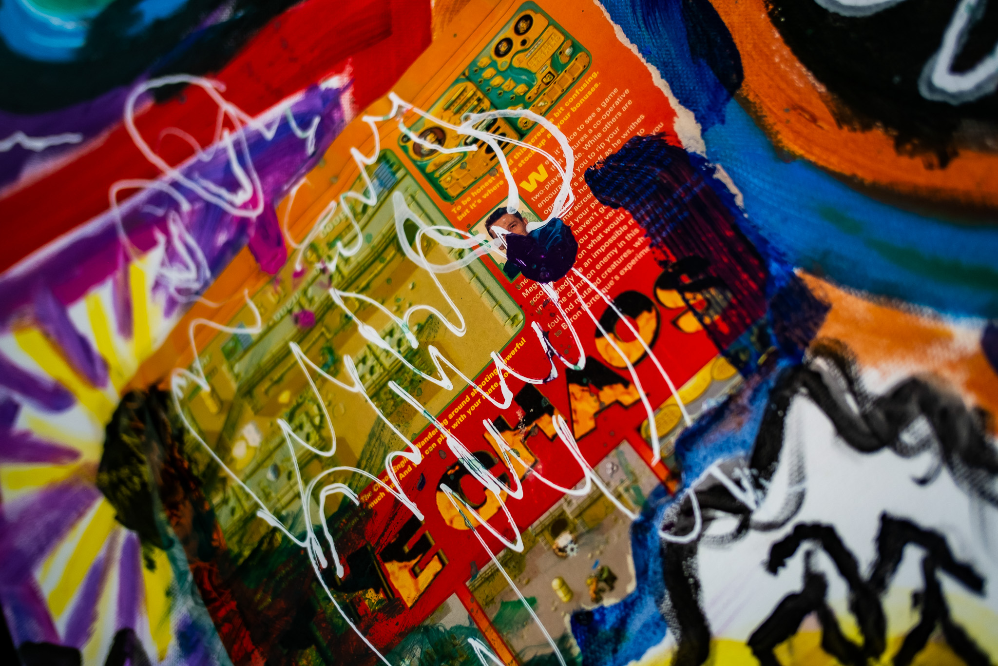 Colourful abstract artwork featuring chaotic layers of vibrant red, orange, yellow, blue, purple, and black shapes, with scattered white scribbles and comic-style illustrations partially visible in the background.
