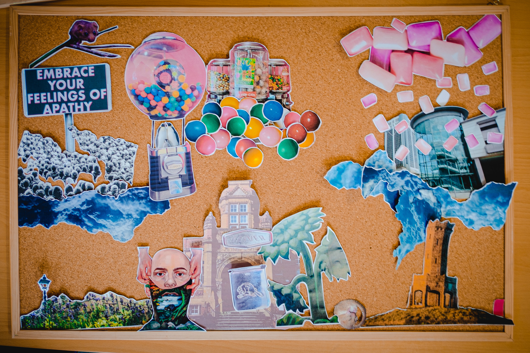A corkboard displays a collage of cut-out images: sweets, colourful gumballs, buildings, clouds, and a crowd. A prominent sign reads “Embrace Your Feelings of Apathy.” There are scattered pieces of pink chewing gum and a person in a mask.