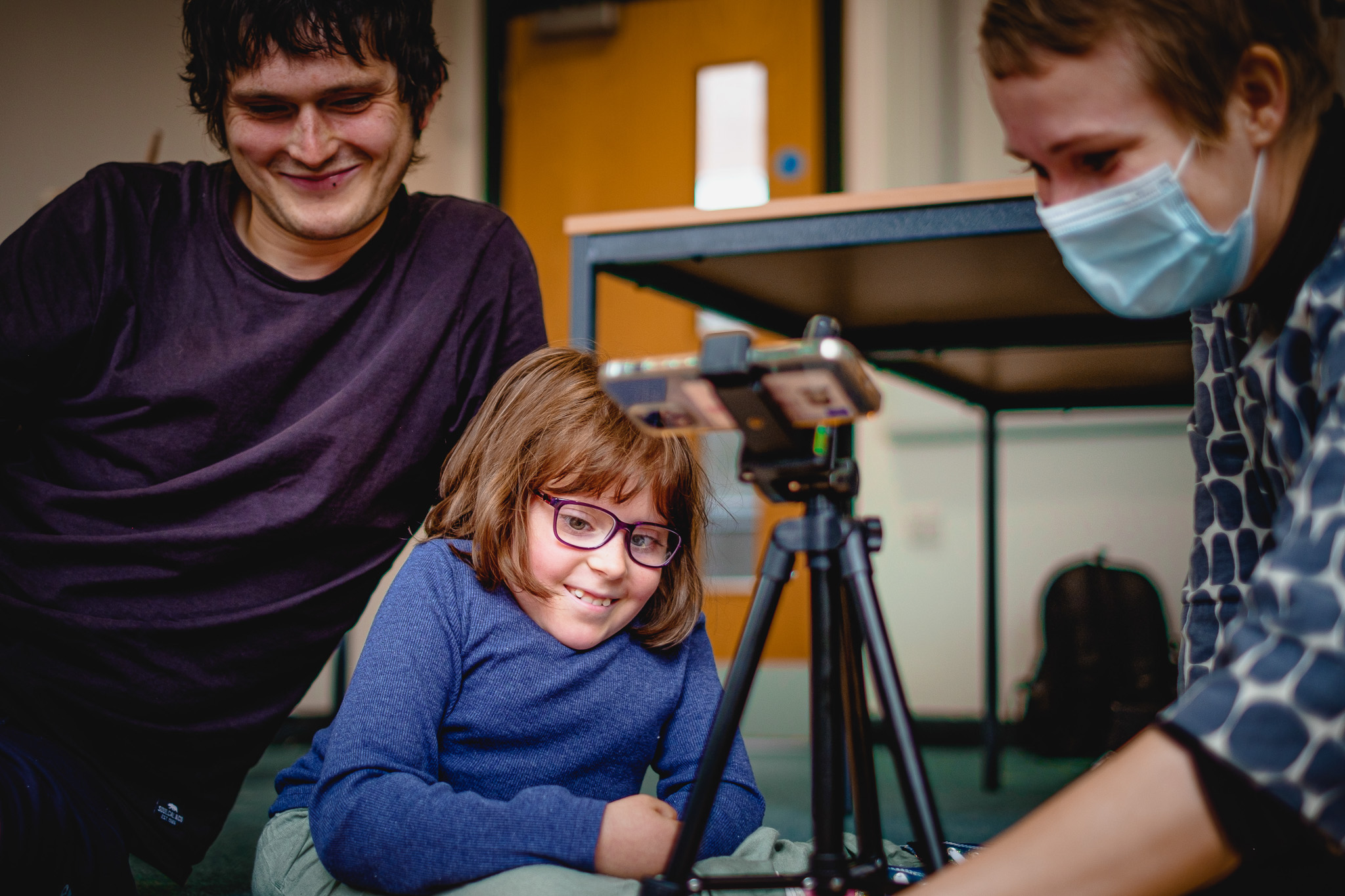 A smiling girl with glasses sits on the floor between two adults, one wearing a mask, gathered around a phone on a tripod.