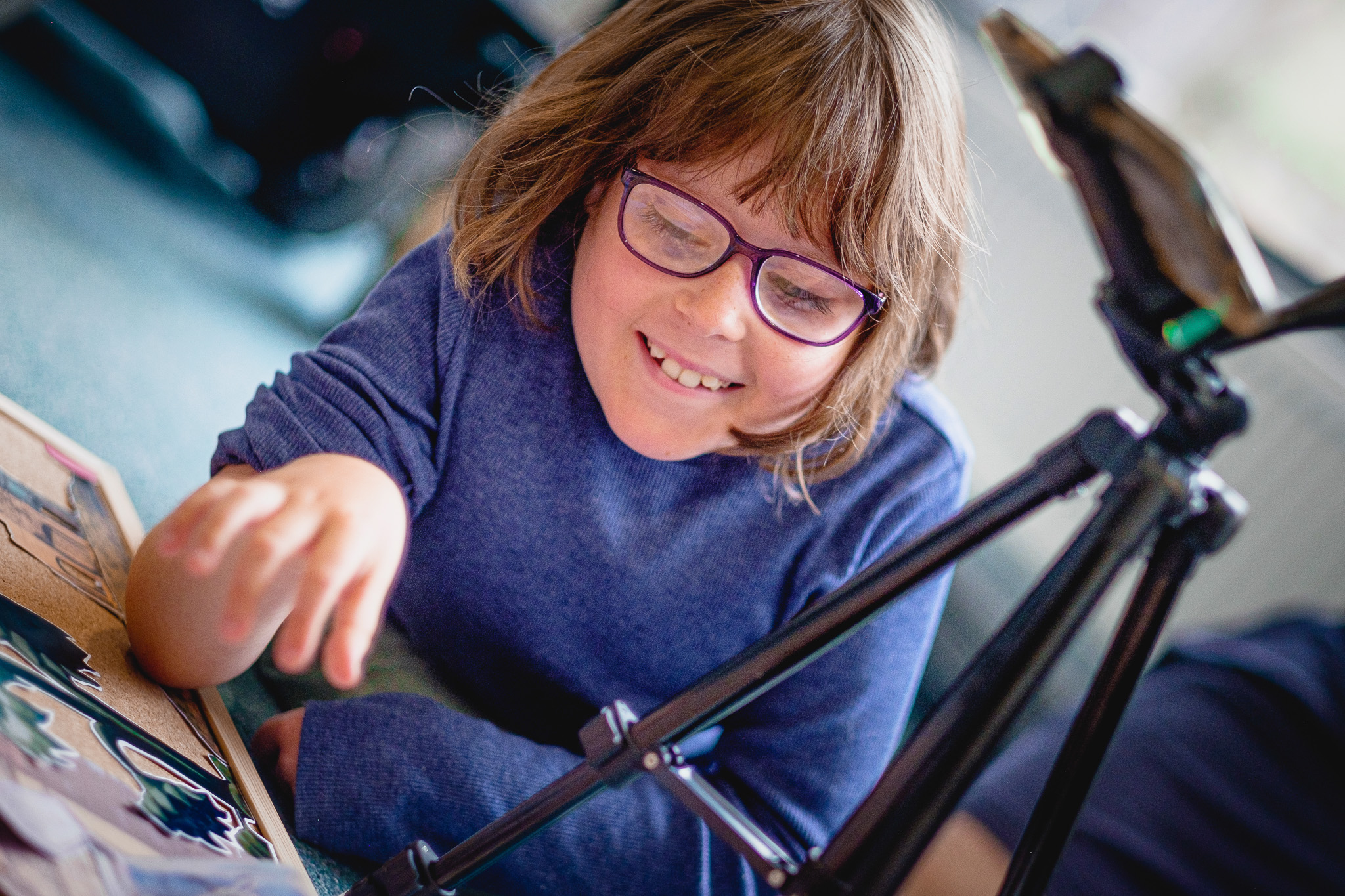 Smiling child with glasses in a blue shirt, pointing at artwork floor, next to a tripod.